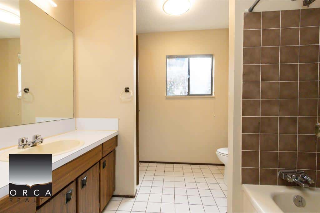 Clean and functional bathroom interior featuring a vanity with a large mirror, a bathtub with brown tiles, and a toilet, perfect for a residential property listing by Orca Realty Inc.
