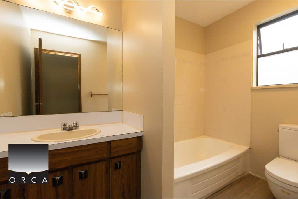 Bright bathroom with a single sink vanity, large mirror, bathtub, and toilet, featuring neutral tones and natural light, ideal for modern residential homes.