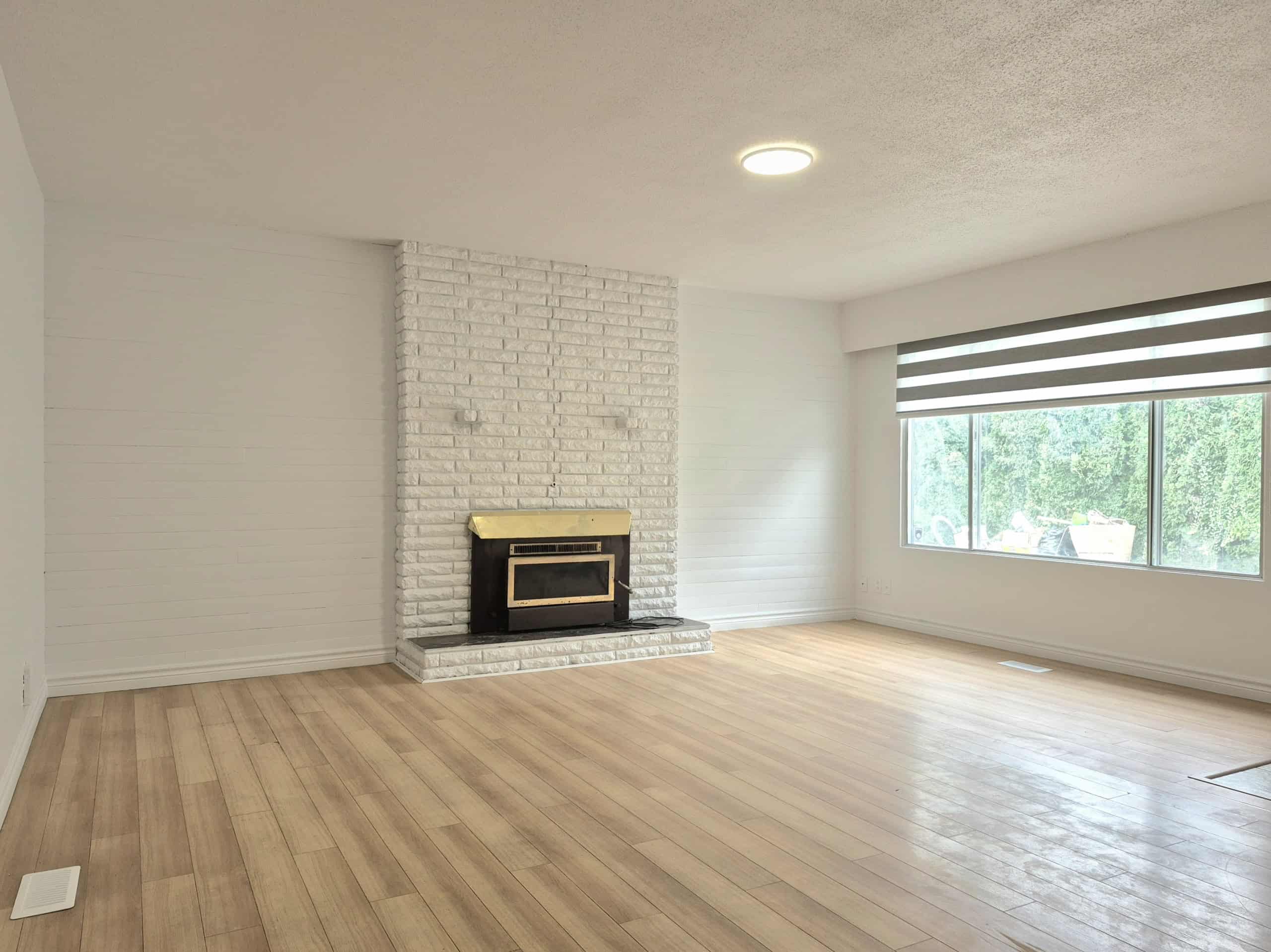 20250920_141124 Spacious living room with white brick fireplace, large window with modern shades, light wood flooring, and bright natural light in a residential property.