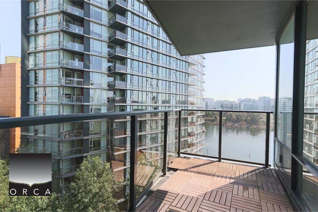 Spacious balcony with glass railings overlooking a river and modern high-rise apartments, ideal for urban living and waterfront views in Vancouver.
