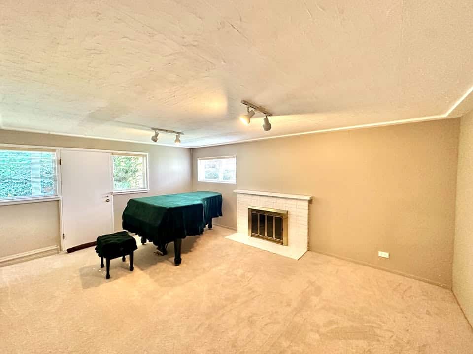 Spacious beige living room with fireplace, windows, and a covered piano, ideal for family gatherings and relaxing, showcasing comfortable home interior decor.