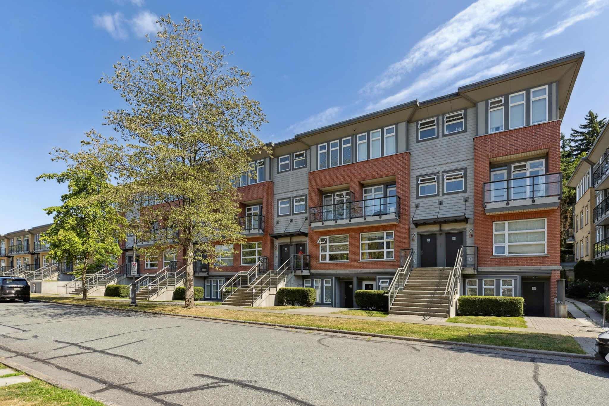 Modern multi-family residential building with brick and grey siding, featuring balconies and outdoor stairs, surrounded by green trees, in a vibrant urban neighbourhood.