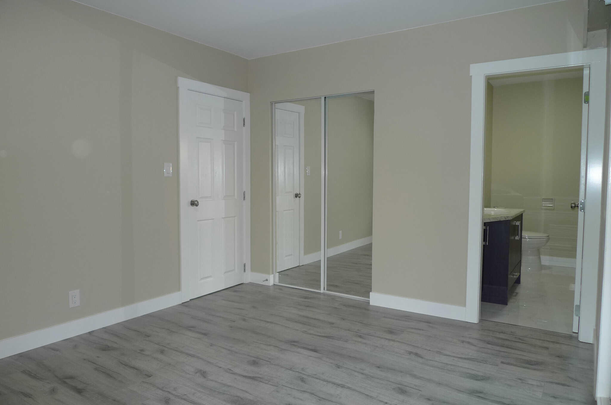 Bright empty bedroom with mirrored closet doors and attached bathroom, showcasing modern interior design, laminate flooring, and neutral wall colors for Ontario homebuyers.