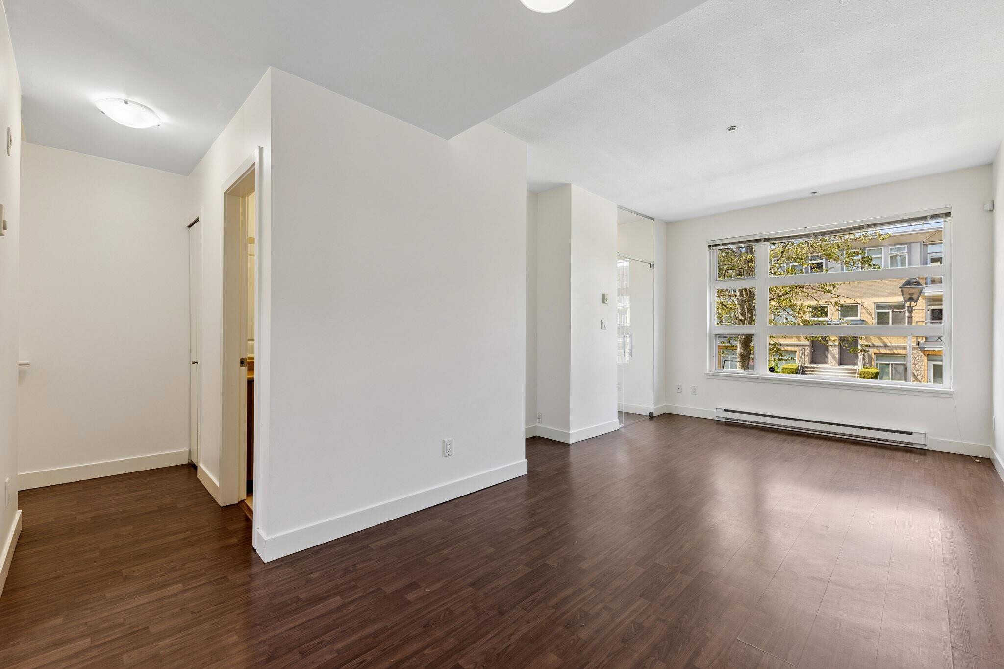 Bright living room with large window, hardwood flooring, and white walls, showcasing a modern condo for sale at Orca Realty Inc. in Vancouver.