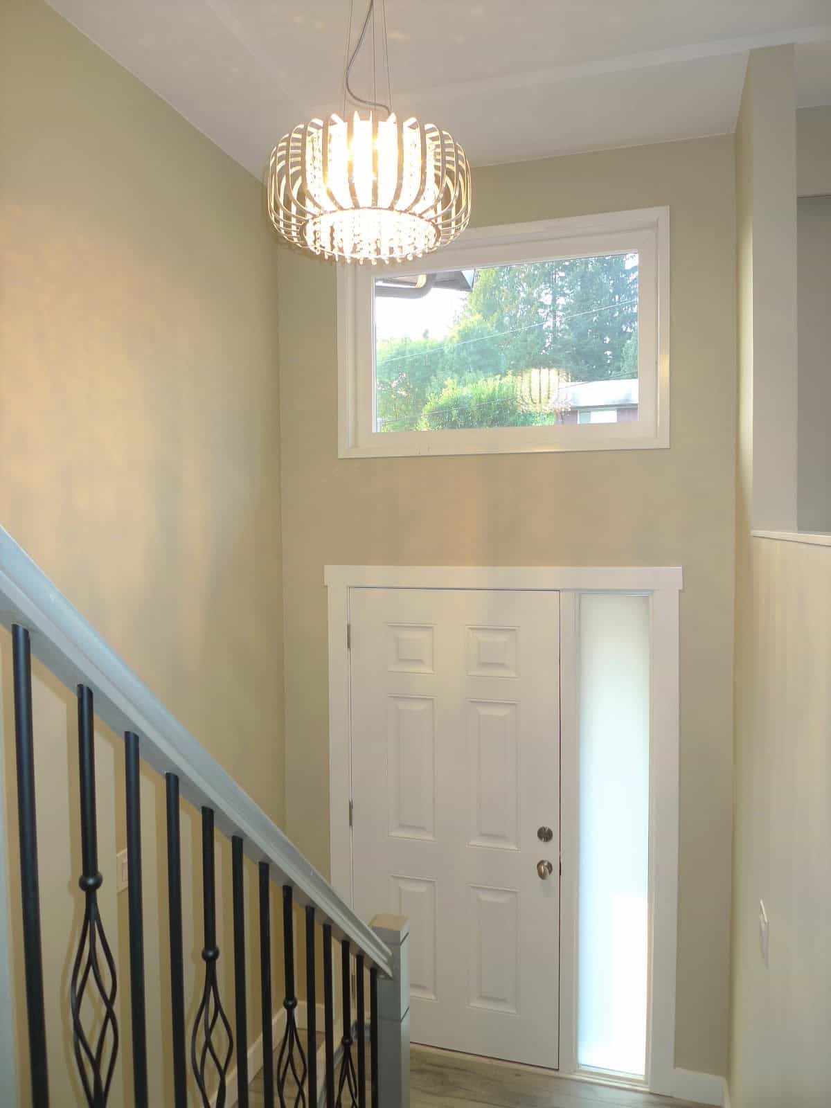 Bright modern interior with a staircase, elegant chandelier, high window, and neutral wall colours, showcasing the inviting entrance of a residential property listed by Orca Realty Inc.