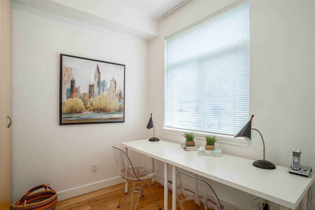 Modern home office with white walls, large window with blinds, minimal decor, and a sleek white desk featuring plants, lamps, and a landline phone for comfortable work-from-home setup.
