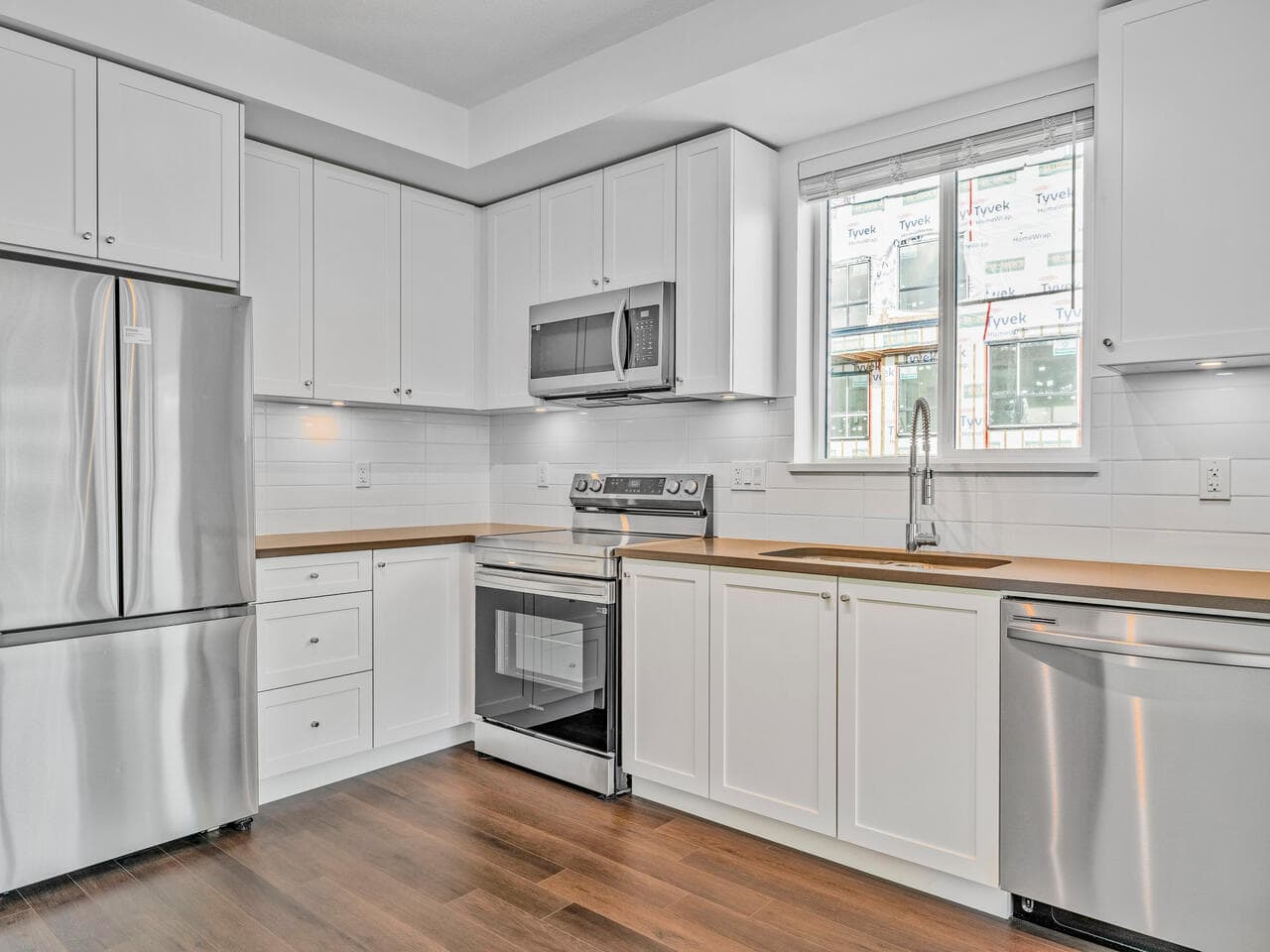Modern white kitchen with stainless steel appliances, wooden countertops, and a large window in a new condo. Featuring a microwave, oven, refrigerator, stove, and dishwasher, perfect for contemporary living.