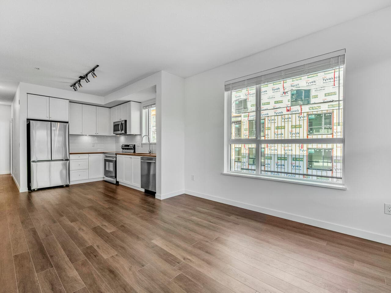 Modern kitchen and living space in a new condo apartment, with white cabinets, stainless steel appliances, large window, hardwood flooring, and fresh interior design at Orca Realty Inc.