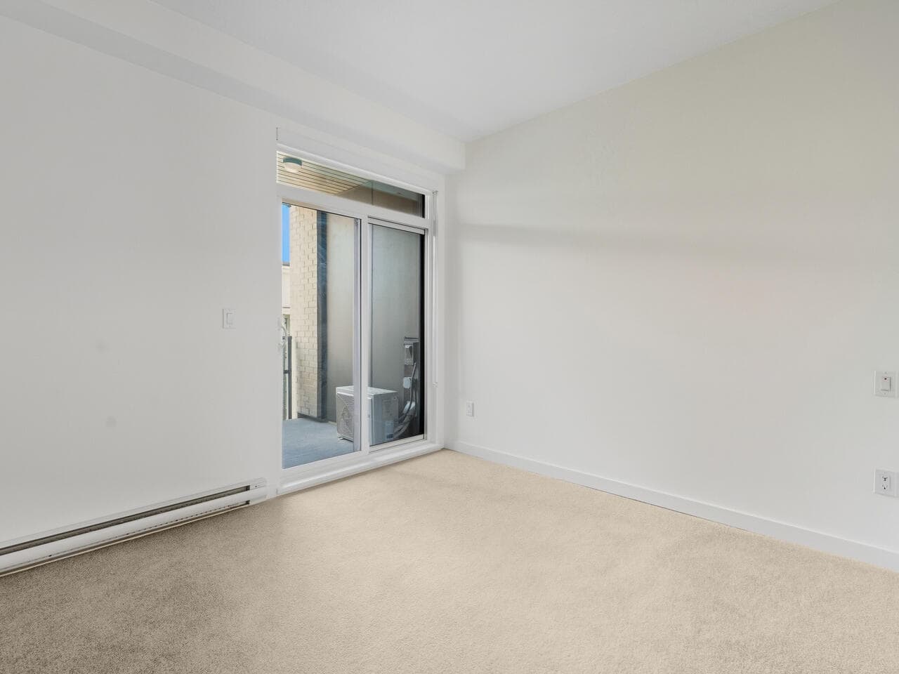 Bright empty condo room with sliding glass door leading to balcony, beige carpet flooring, ideal for Vancouver real estate listings and residential property marketing.