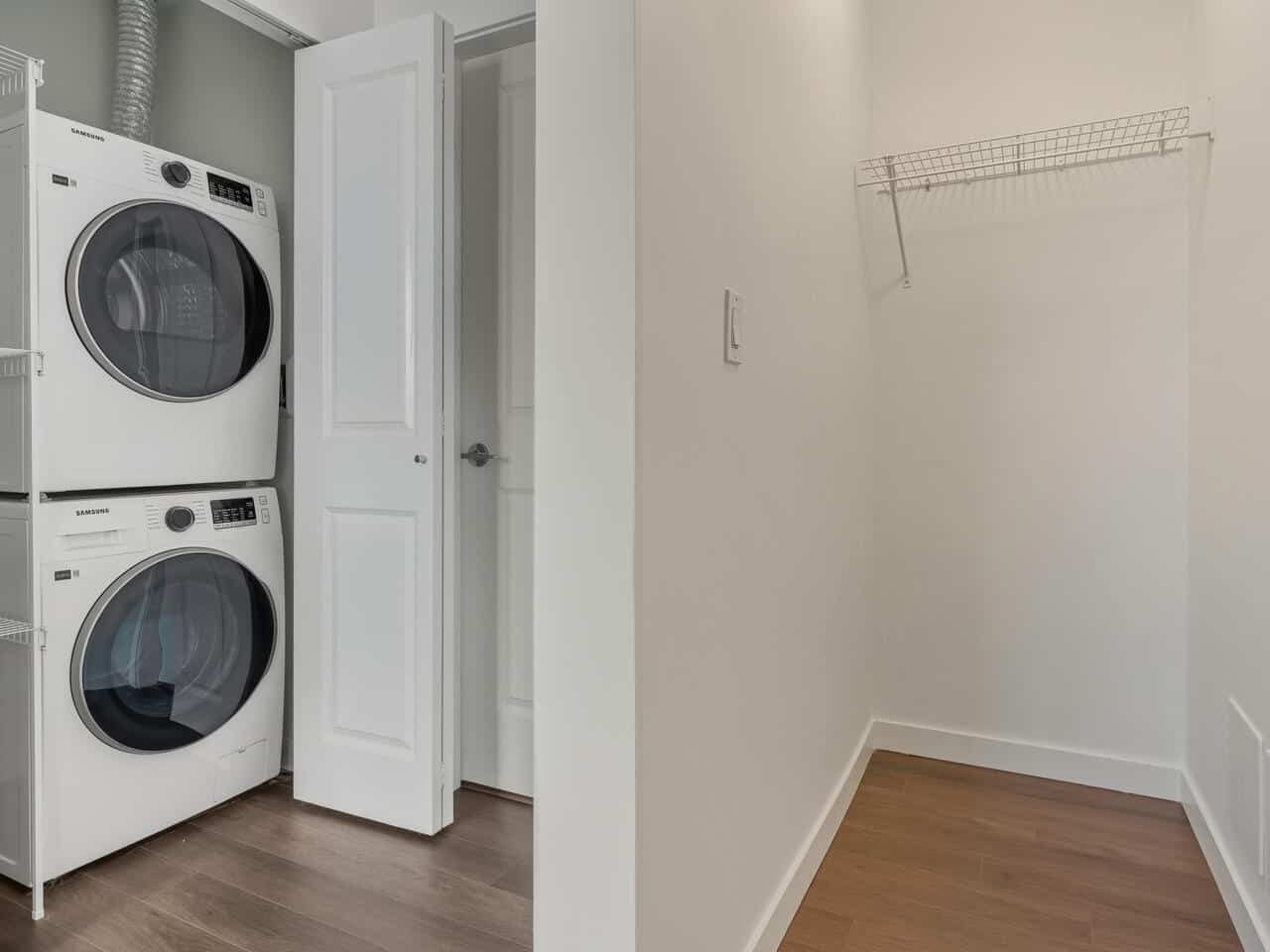Stacked front-load washer and dryer in a white laundry room with hardwood flooring, a small wire shelf on the wall, and a closet door, showcasing an organized, modern laundry space.