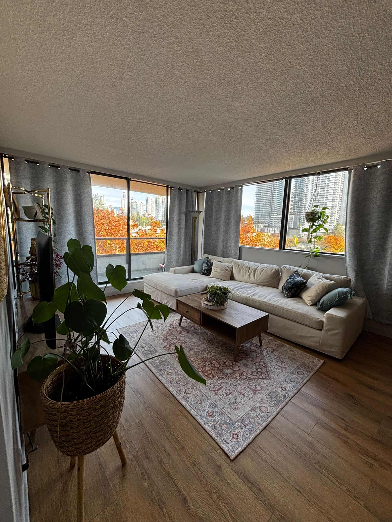Bright living room with large windows showcasing city skyline and autumn trees, featuring a white sectional sofa, wooden coffee table, indoor plants, and cozy curtains.