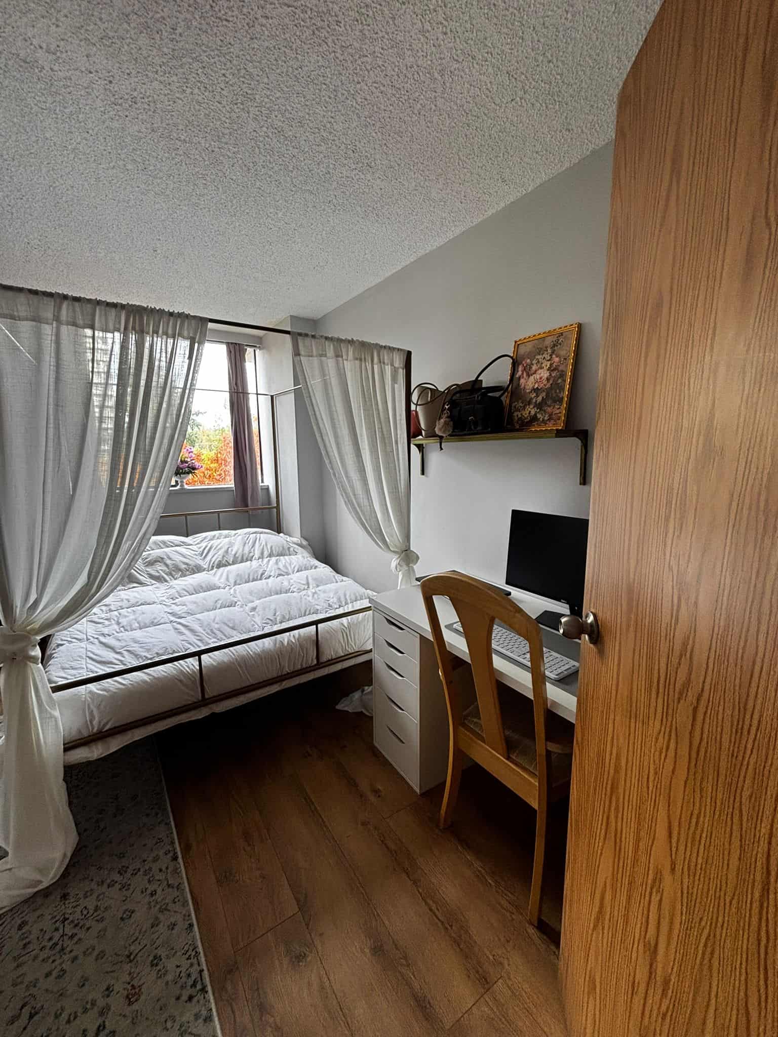 Cozy bedroom with white curtains, a bed, and a small workspace featuring a computer and wooden chair, perfect for comfortable living and home office setup.