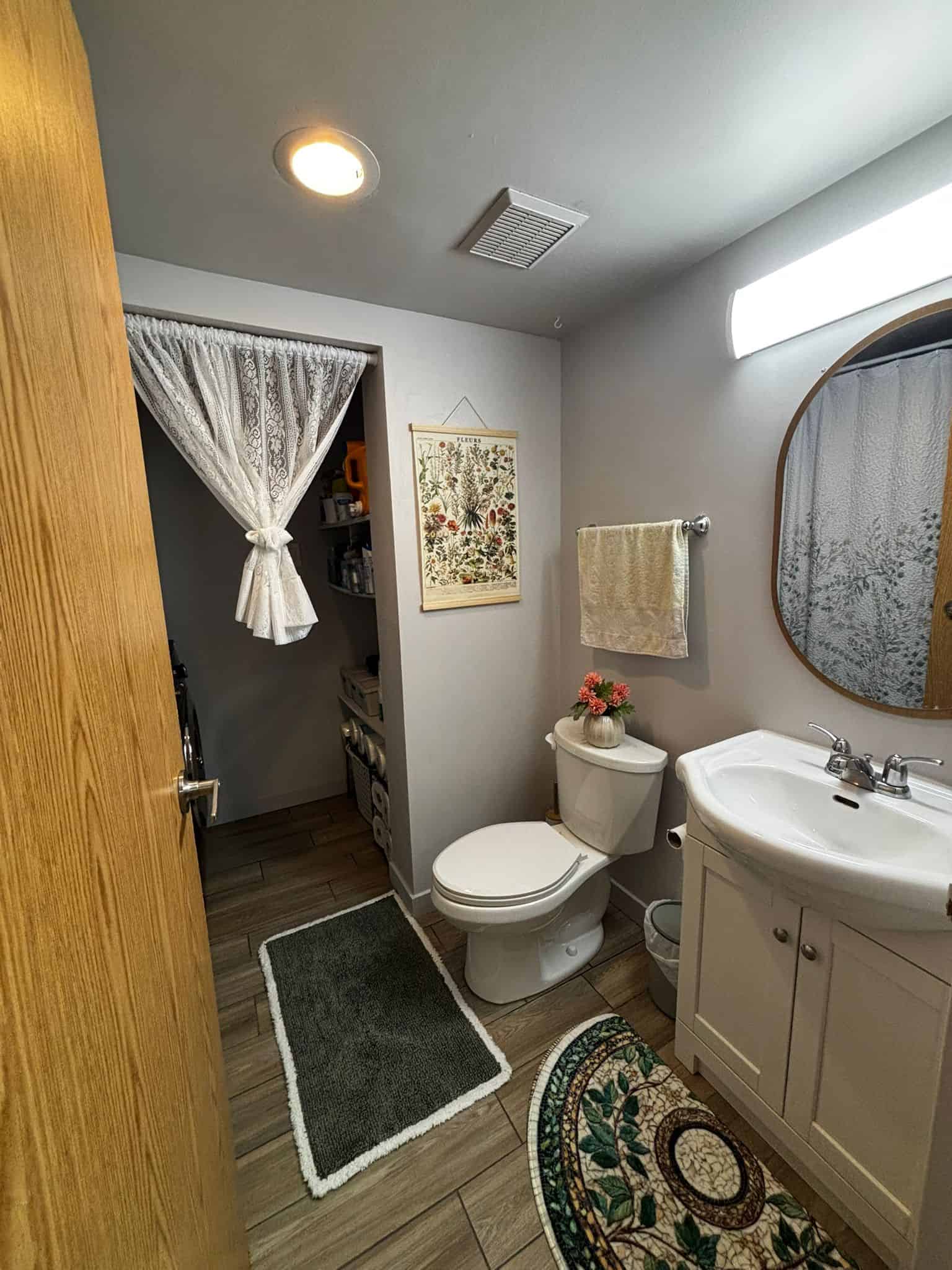 Cozy bathroom with white fixtures, decorative rugs, and a botanical wall art, featuring a vanity with storage, a mirror, and a small laundry or storage nook with lace curtains, perfect for a home in British Columbia.