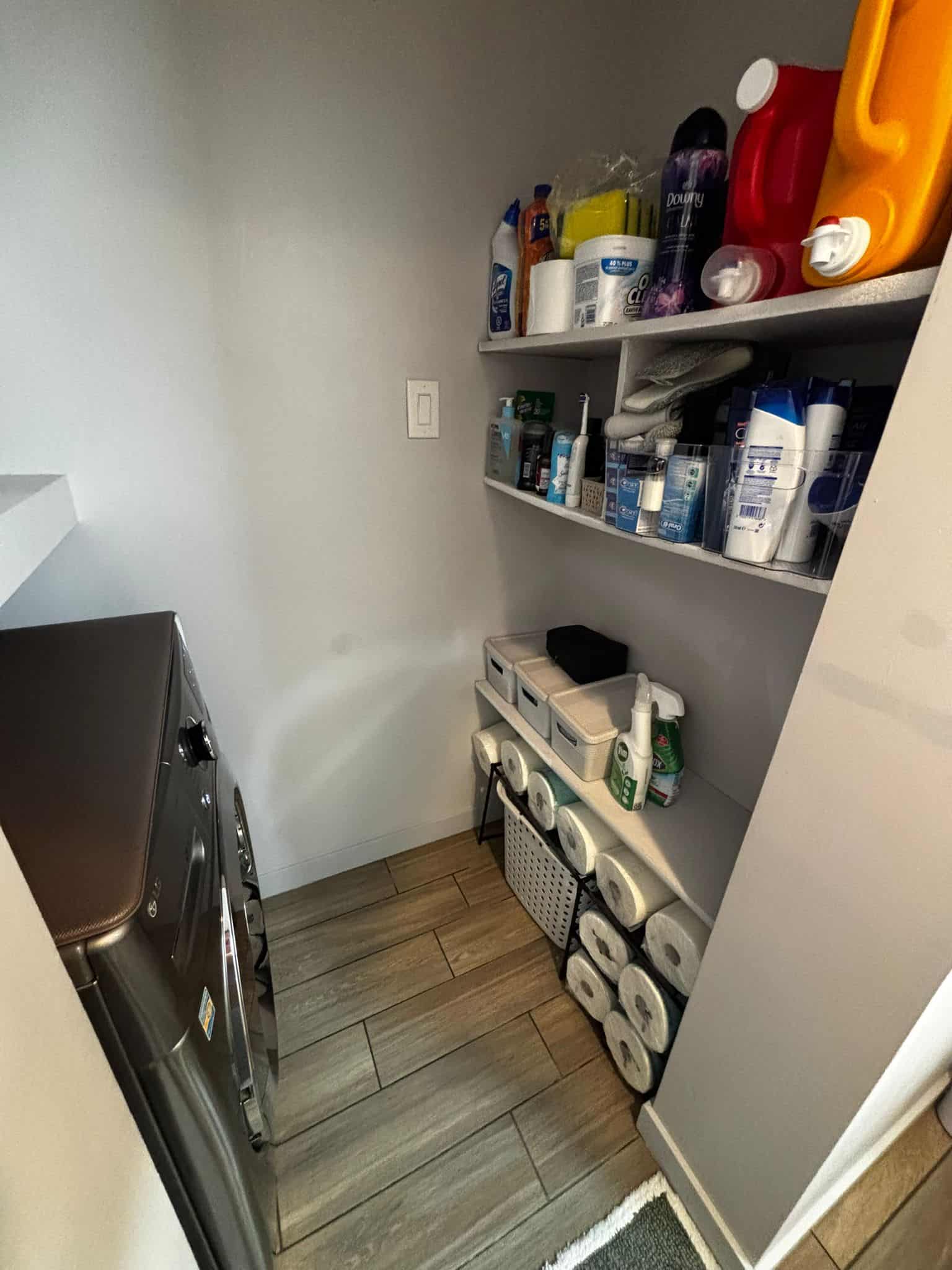 Stainless steel washing machine in a laundry room with storage shelves containing cleaning supplies, toiletries, and paper towels, showcasing organized utility space at Orca Realty Inc.