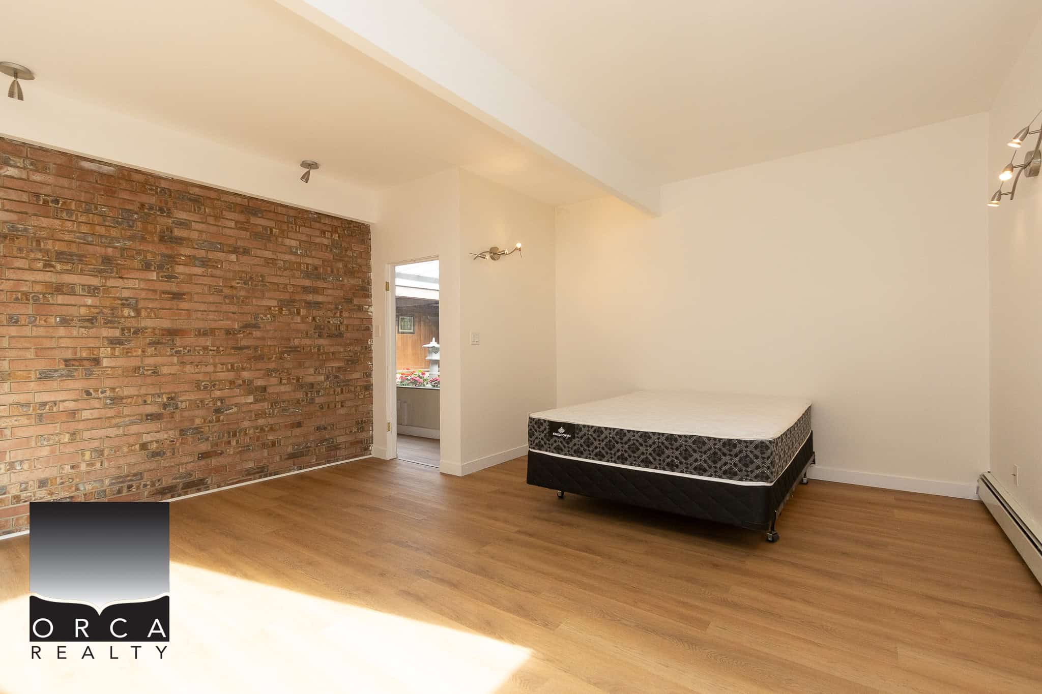 Bright, spacious bedroom with hardwood floors, exposed brick accent wall, and large window providing natural light in a modern rental property listed by Orca Realty Inc.