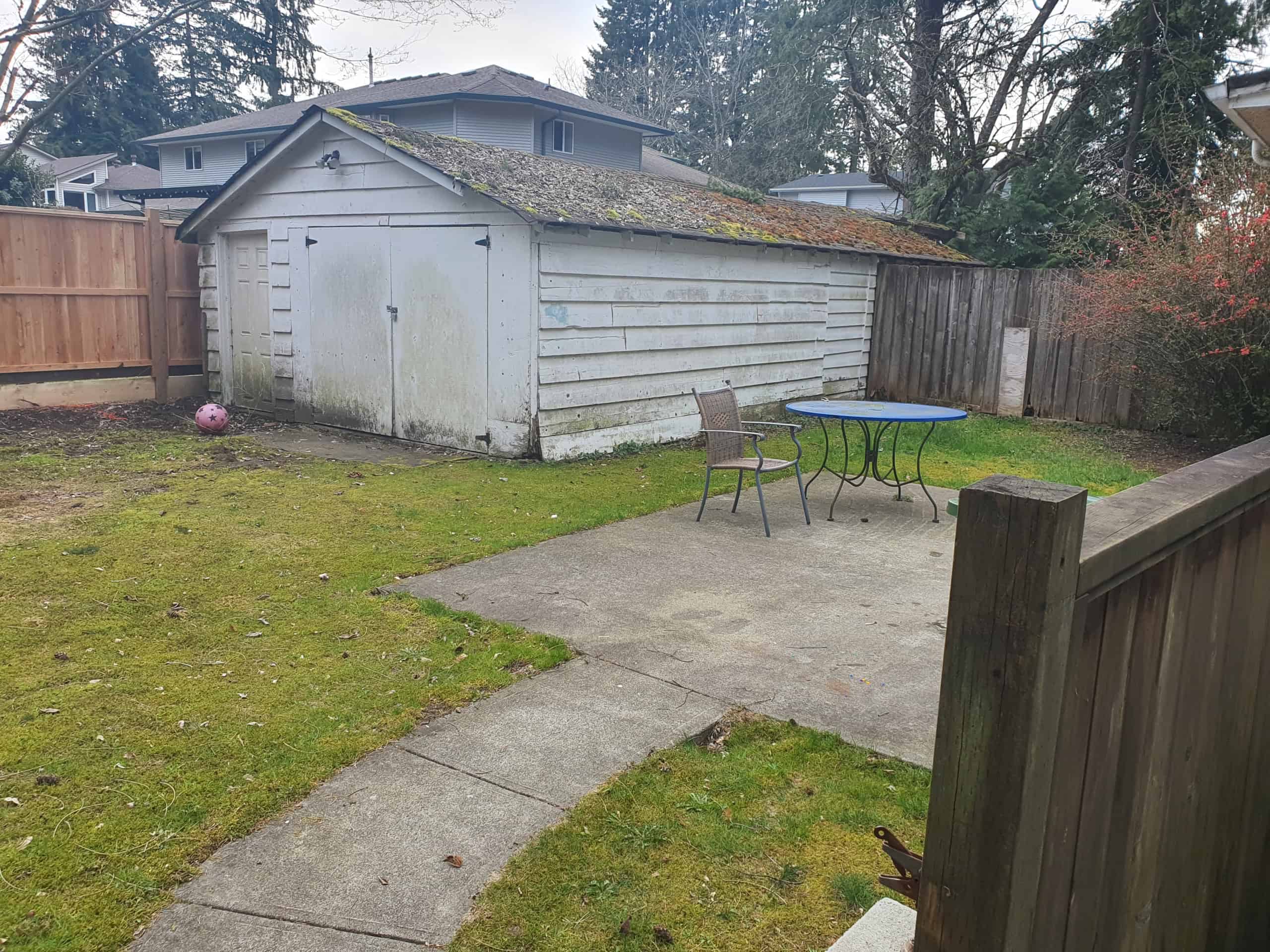 Weathered backyard shed with moss-covered roof in a suburban yard with patio furniture and fencing for privacy, ideal for storage or gardening supplies.