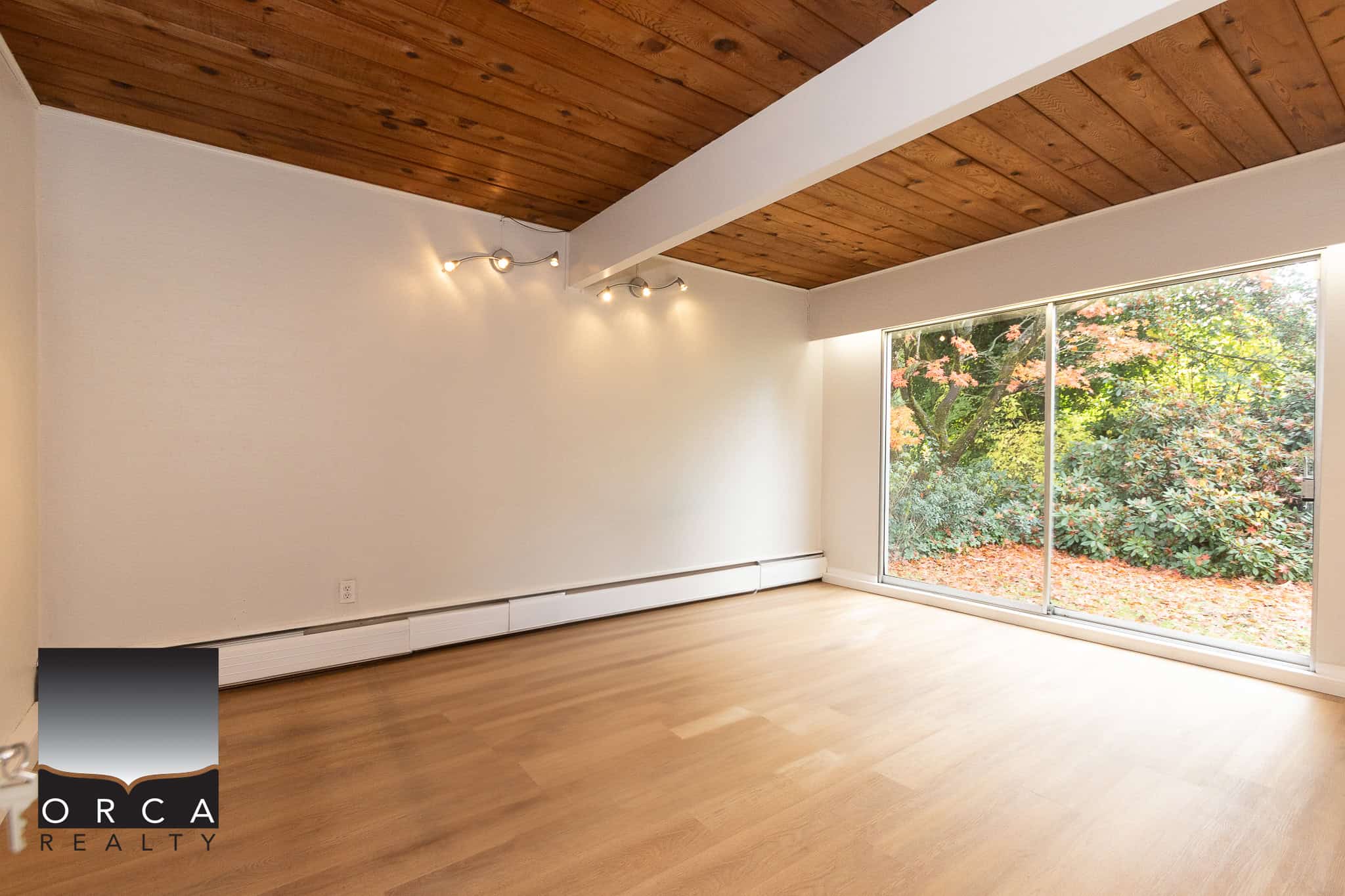Bright and spacious empty living room with large windows showcasing lush greenery, hardwood floors, and a wooden ceiling, perfect for Vancouver and surrounding area property listings.
