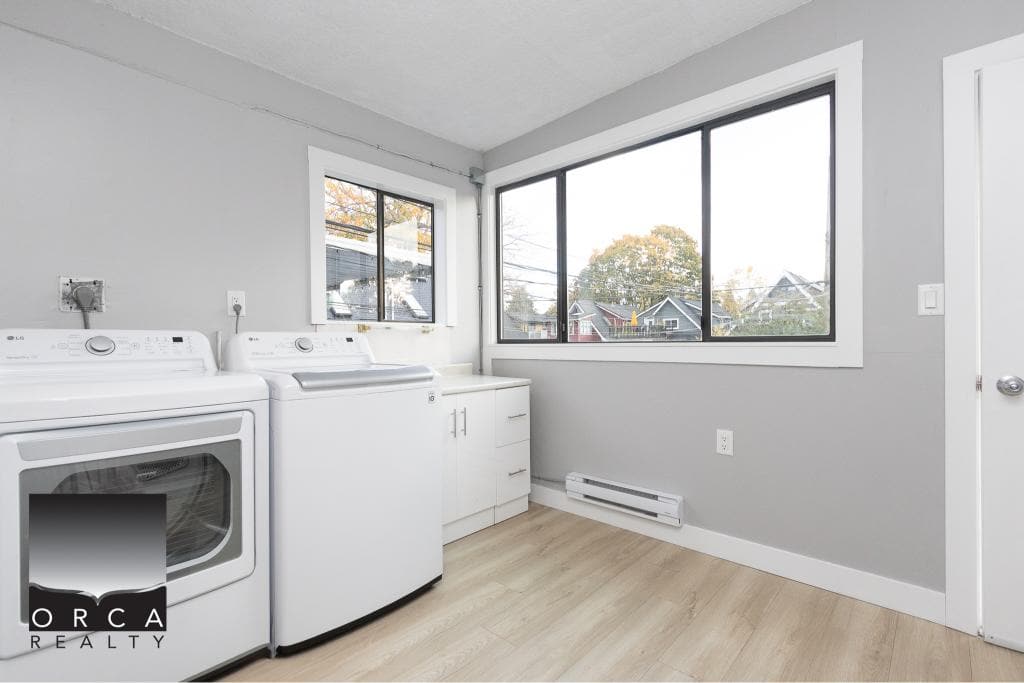 Washer and dryer in a bright laundry room with large windows, modern appliances, light gray walls, and wood flooring, ideal for Vancouver area homes.