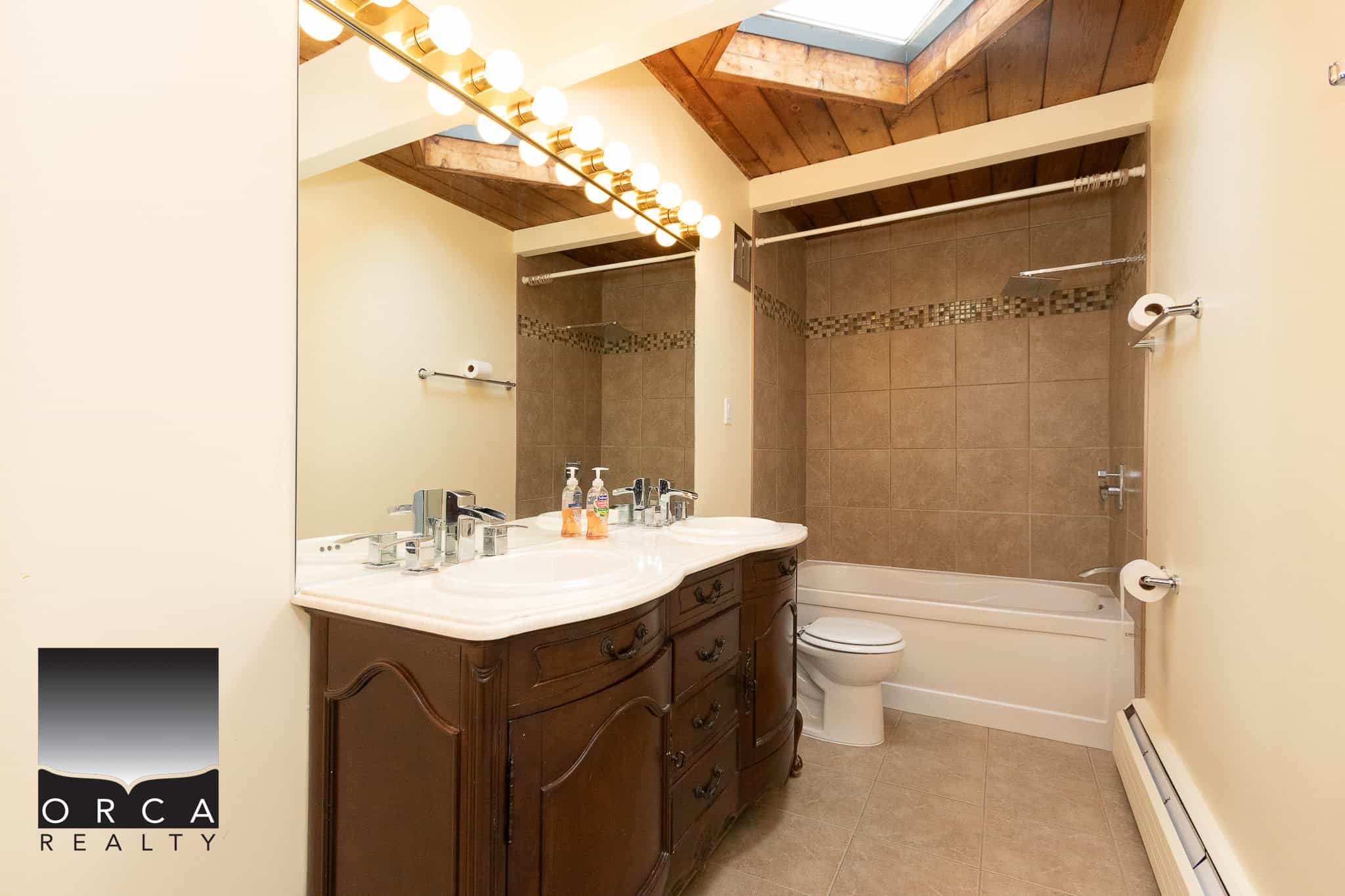 Bright bathroom with double vanity, bathtub, beige tile walls, and illuminated mirror, showcasing modern home features in a property listed by Orca Realty Inc.