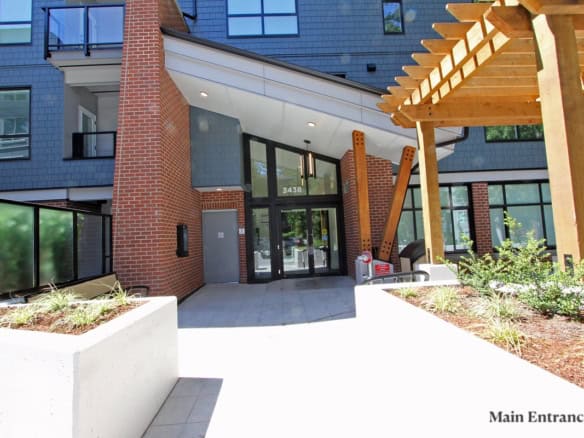 Bright modern apartment building entrance with brick and glass design, landscaped planters, and wooden pergola, showcasing Orca Realty Inc.’s professional property management services in Vancouver.
