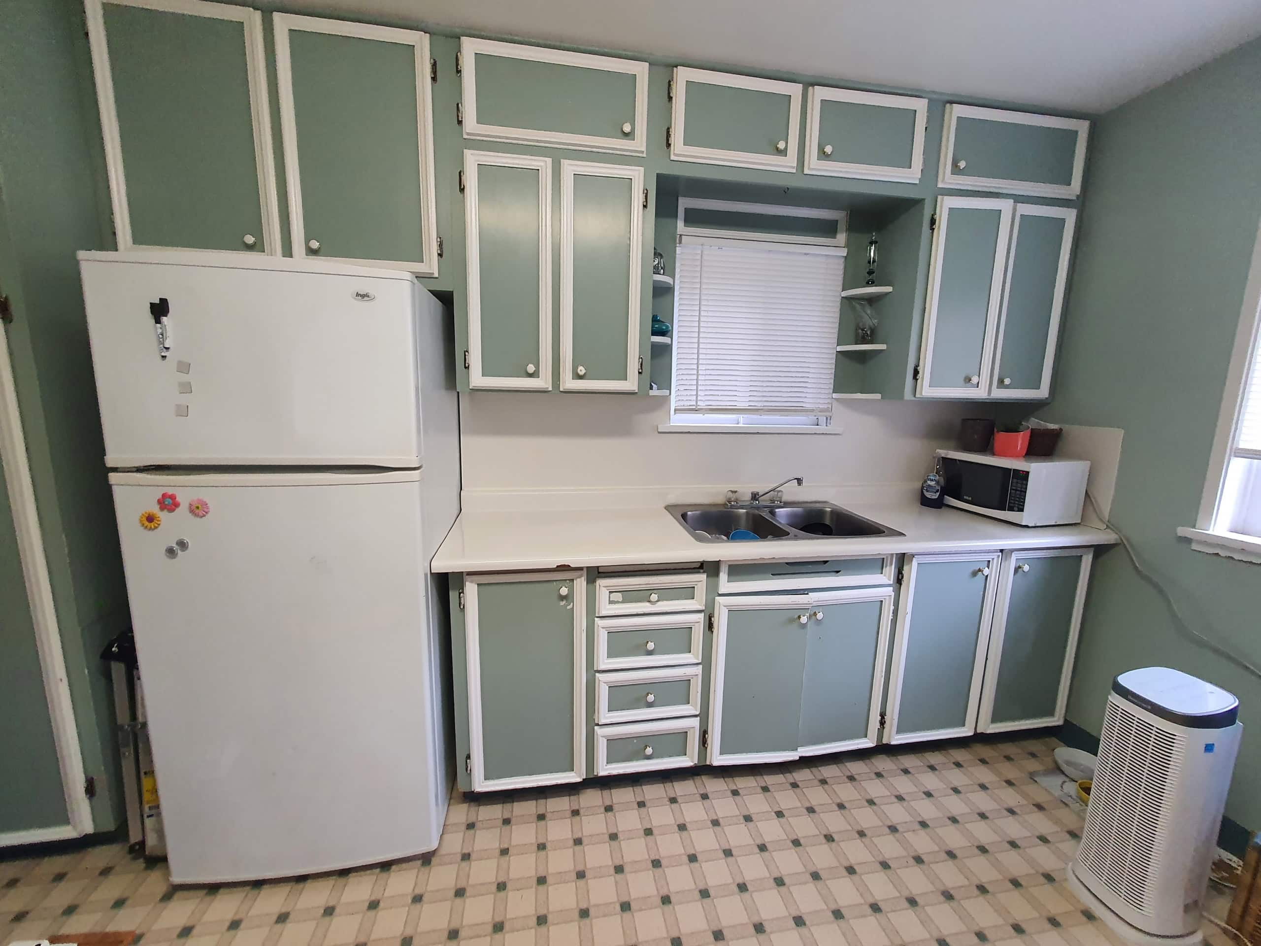 Vintage kitchen with pastel green and white cabinets, compact refrigerator, microwave, and window blinds, perfect for homebuyers seeking classic, functional kitchen spaces.