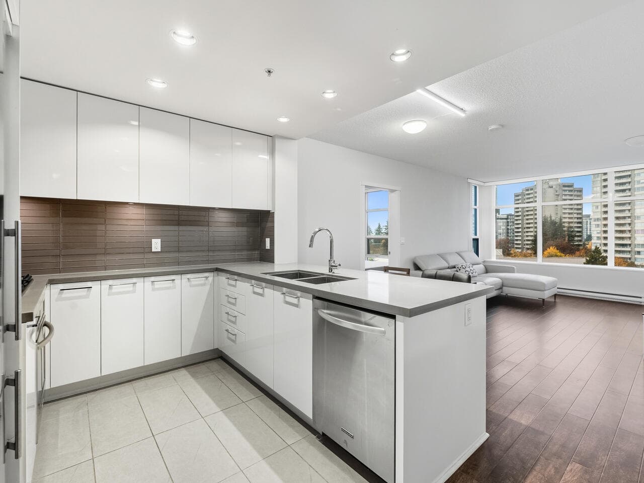 Modern white kitchen with sleek cabinetry and stainless steel appliances in a bright, open-concept living space with large windows and city views.