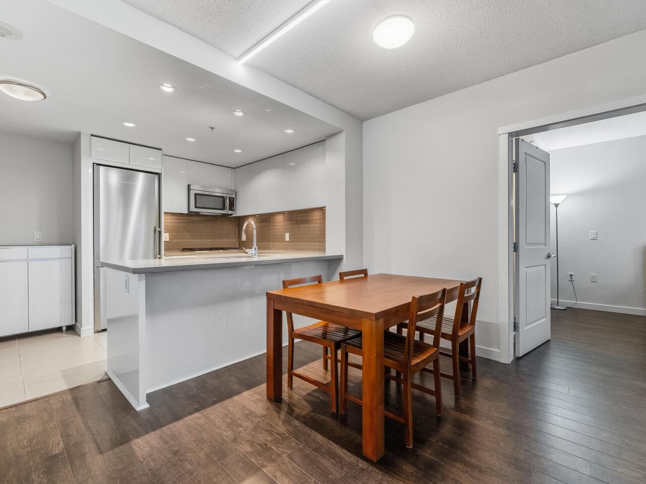 Spacious modern kitchen and dining area in Vancouver apartment, featuring sleek white cabinetry, stainless steel appliances, dark hardwood floors, and a cozy dining table for family meals.