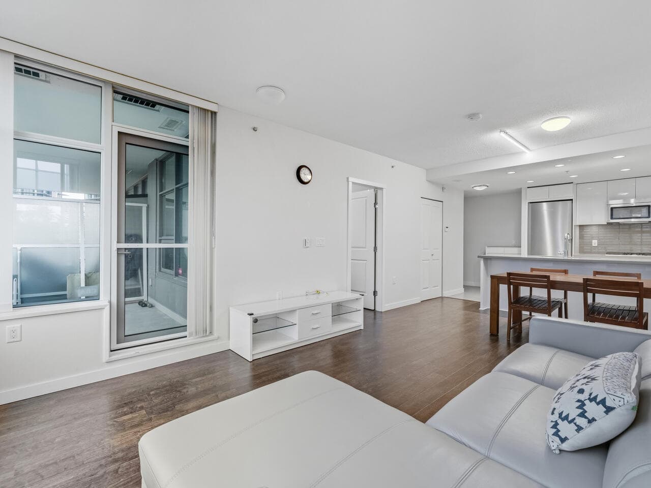 Bright, modern condo living room with hardwood floors, large windows, and a cozy white sofa, featuring an open-concept kitchen and balcony access, ideal for Vancouver real estate enthusiasts.