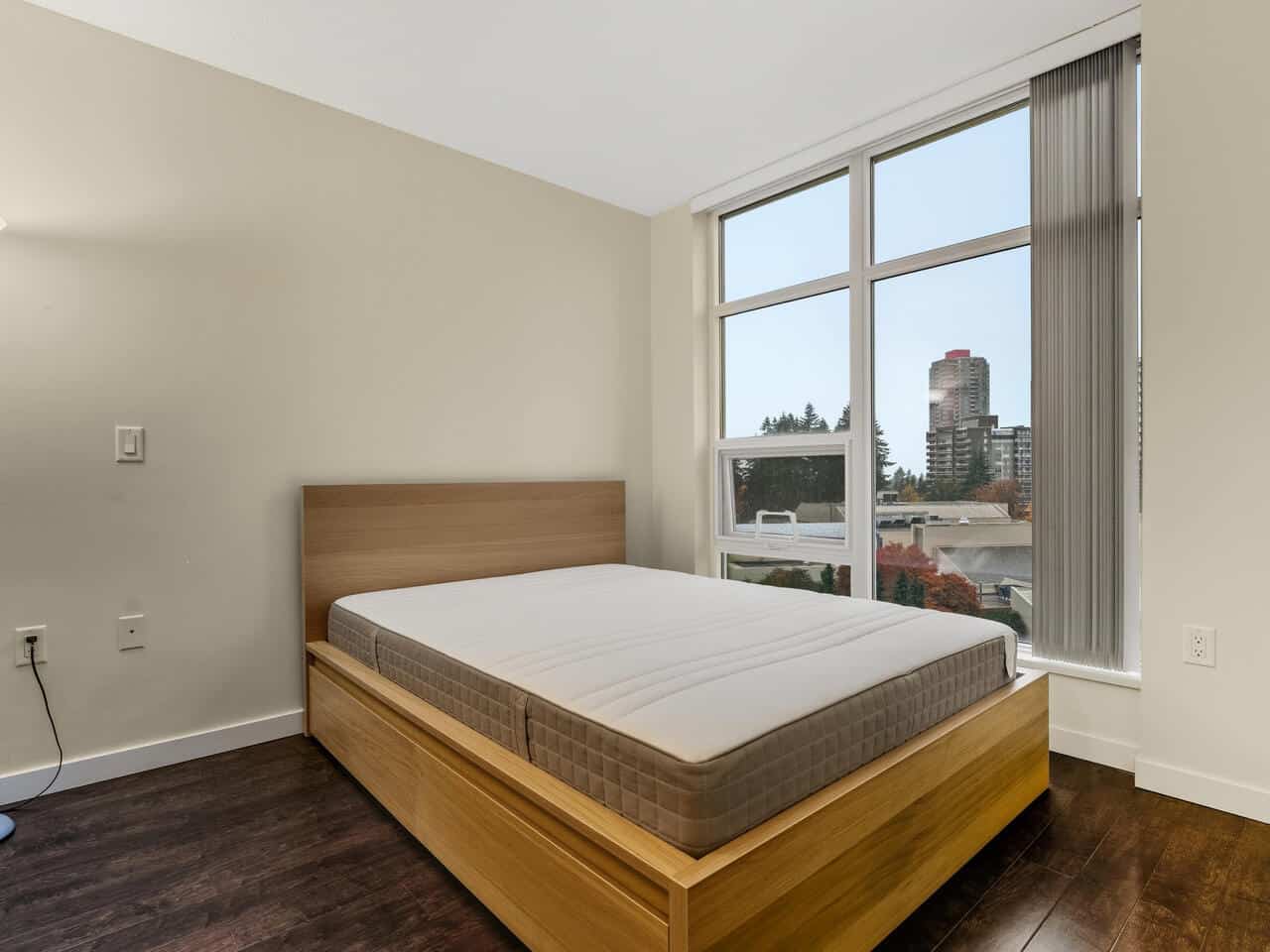Modern bedroom with large windows showcasing city views, oak furniture, and dark wood flooring, perfect for Vancouver-area real estate listings by Orca Realty Inc.