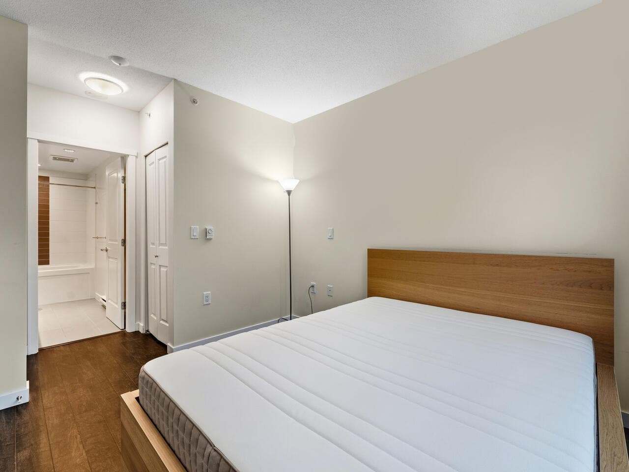 Contemporary bedroom with a wooden headboard, clean white bedding, and minimalist decor, featuring modern lighting and access to an en-suite bathroom – perfect for Vancouver real estate and residential rentals.
