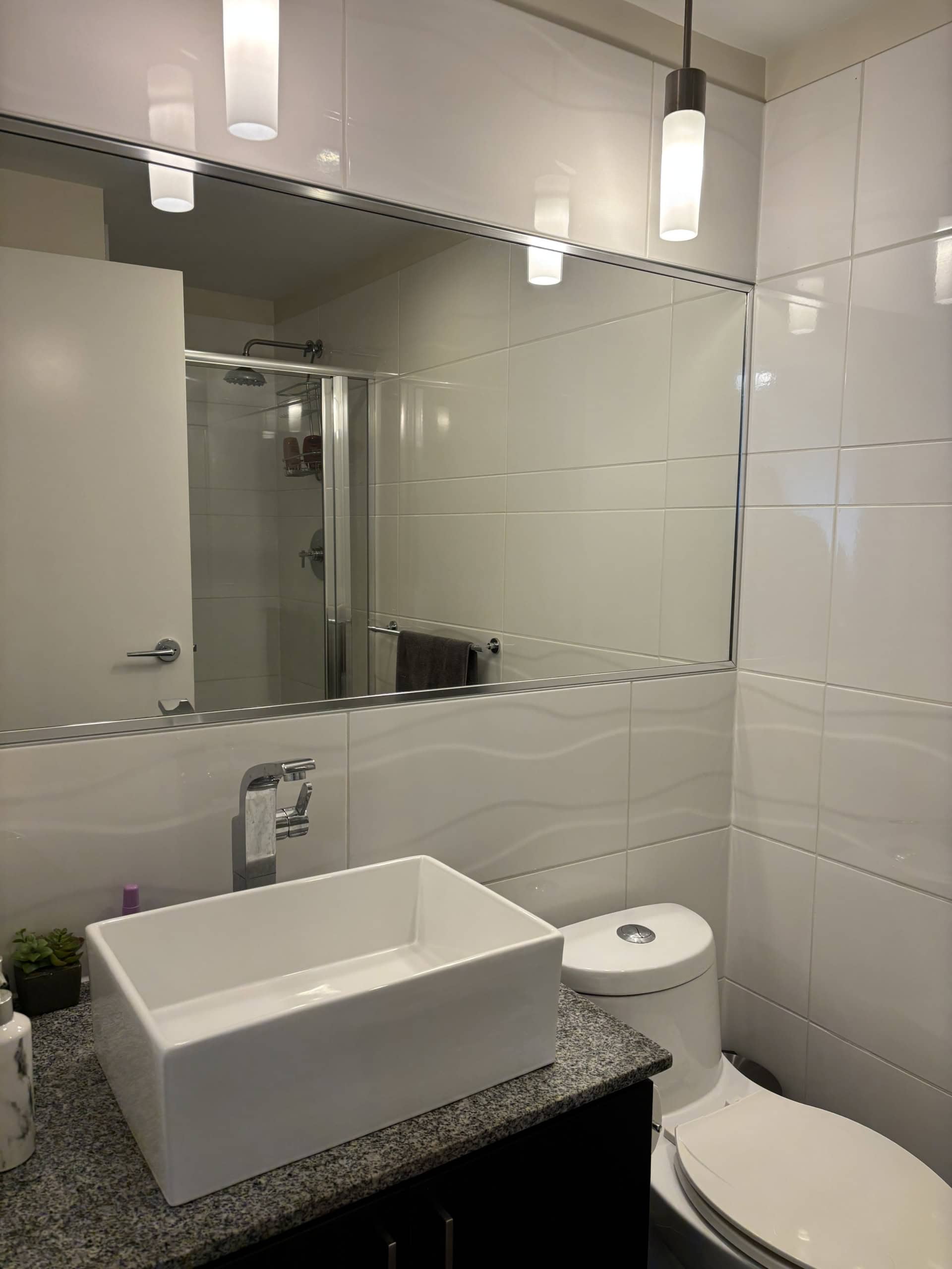 Modern bathroom with white tile walls, sleek vanity with vessel sink, contemporary fixtures, and a large mirror reflecting a shower area with glass door, highlighting stylish, functional residential real estate in Vancouver.