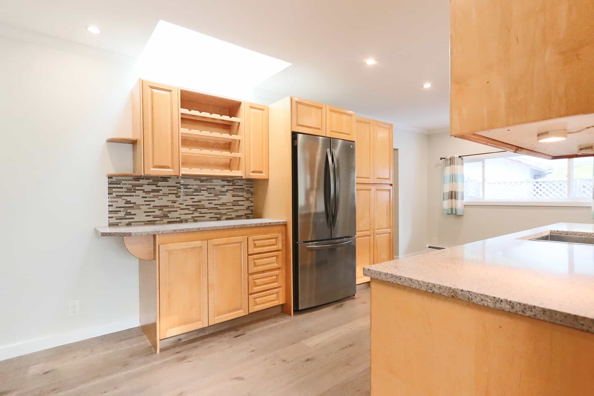 Bright modern kitchen with natural wood cabinets, stainless steel refrigerator, granite countertops, mosaic tile backsplash, and ample natural light in a serene home setting.