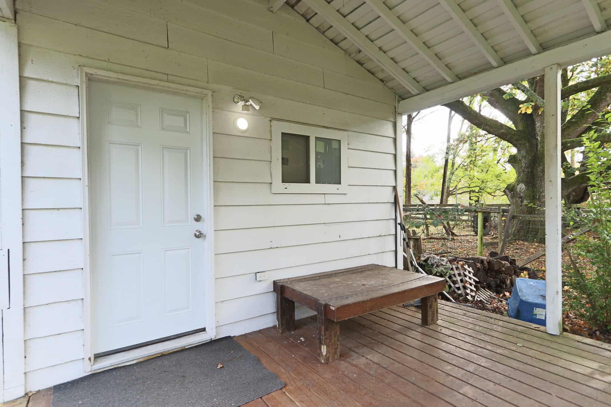 Bright outdoor covered porch area of a residential property, featuring a white wooden door, window, and rustic wooden bench with a natural wooded yard in the background, ideal for outdoor living and relaxation.