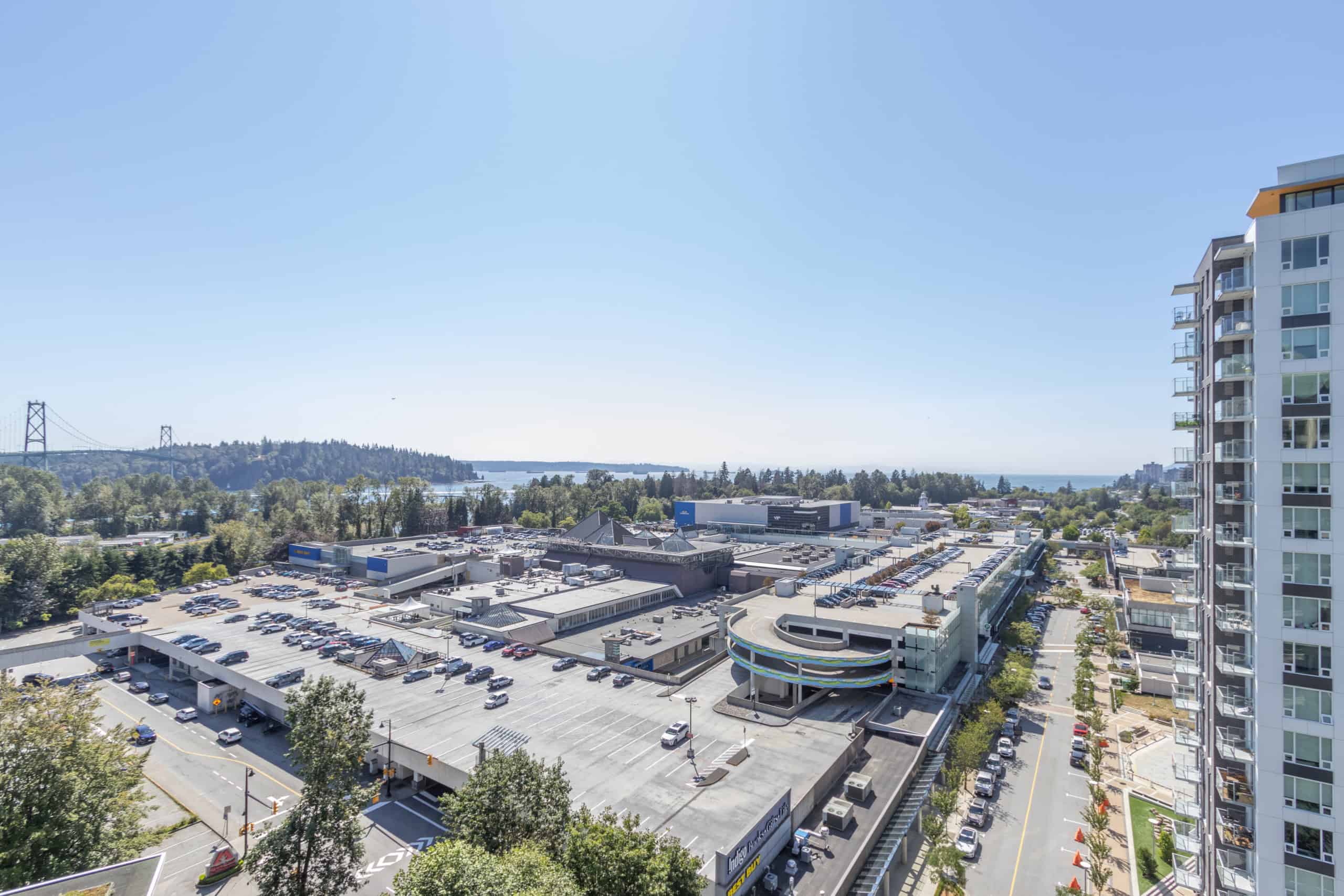 Modern Vancouver waterfront skyline view from high-rise condo, featuring downtown architecture, shopping mall, lush greenery, and distant mountains with a clear blue sky.