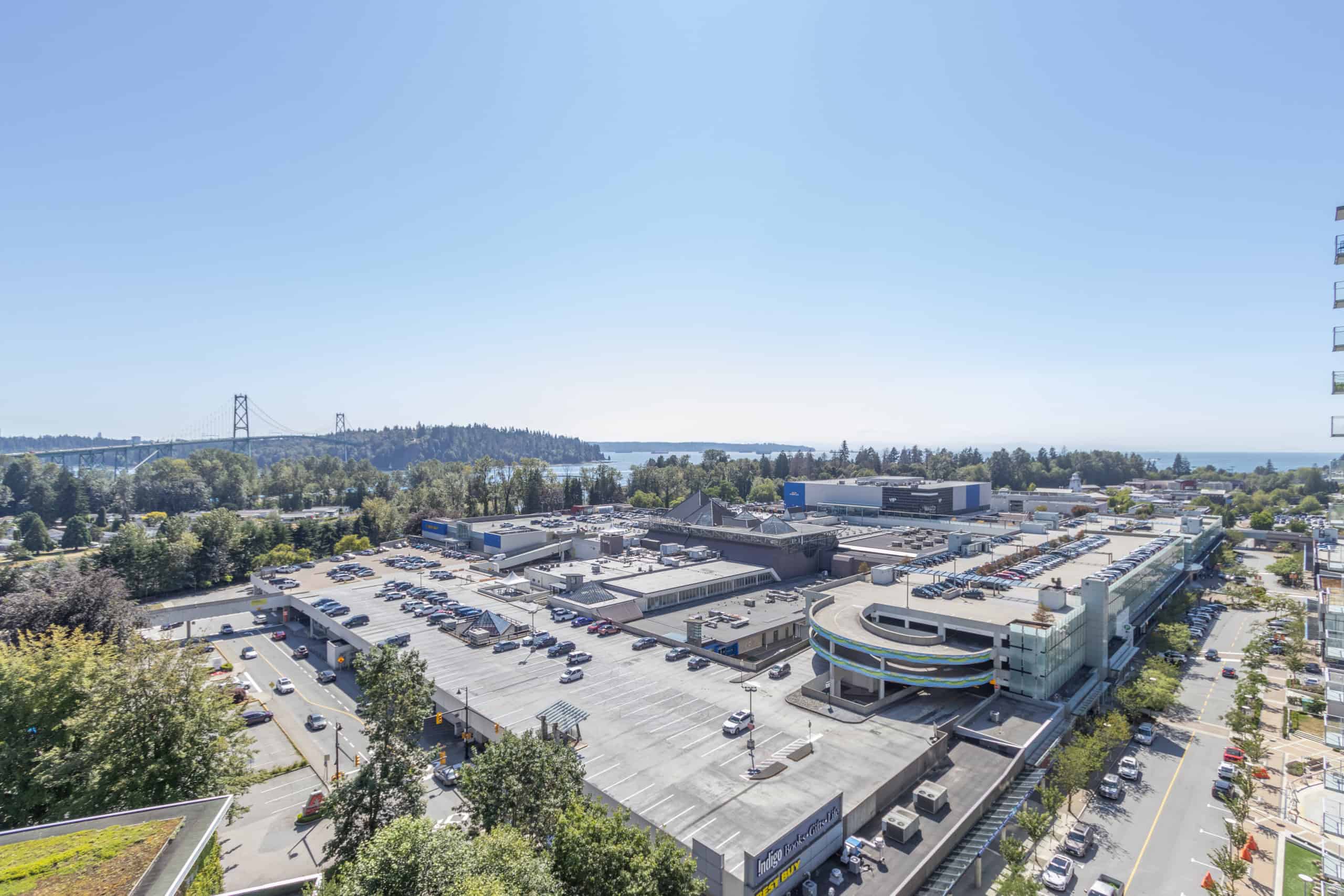 Aerial view of a commercial shopping center with parking lot, surrounded by lush green trees, with a distant view of a bridge and water body, showcasing urban development and scenic landscape in British Columbia.