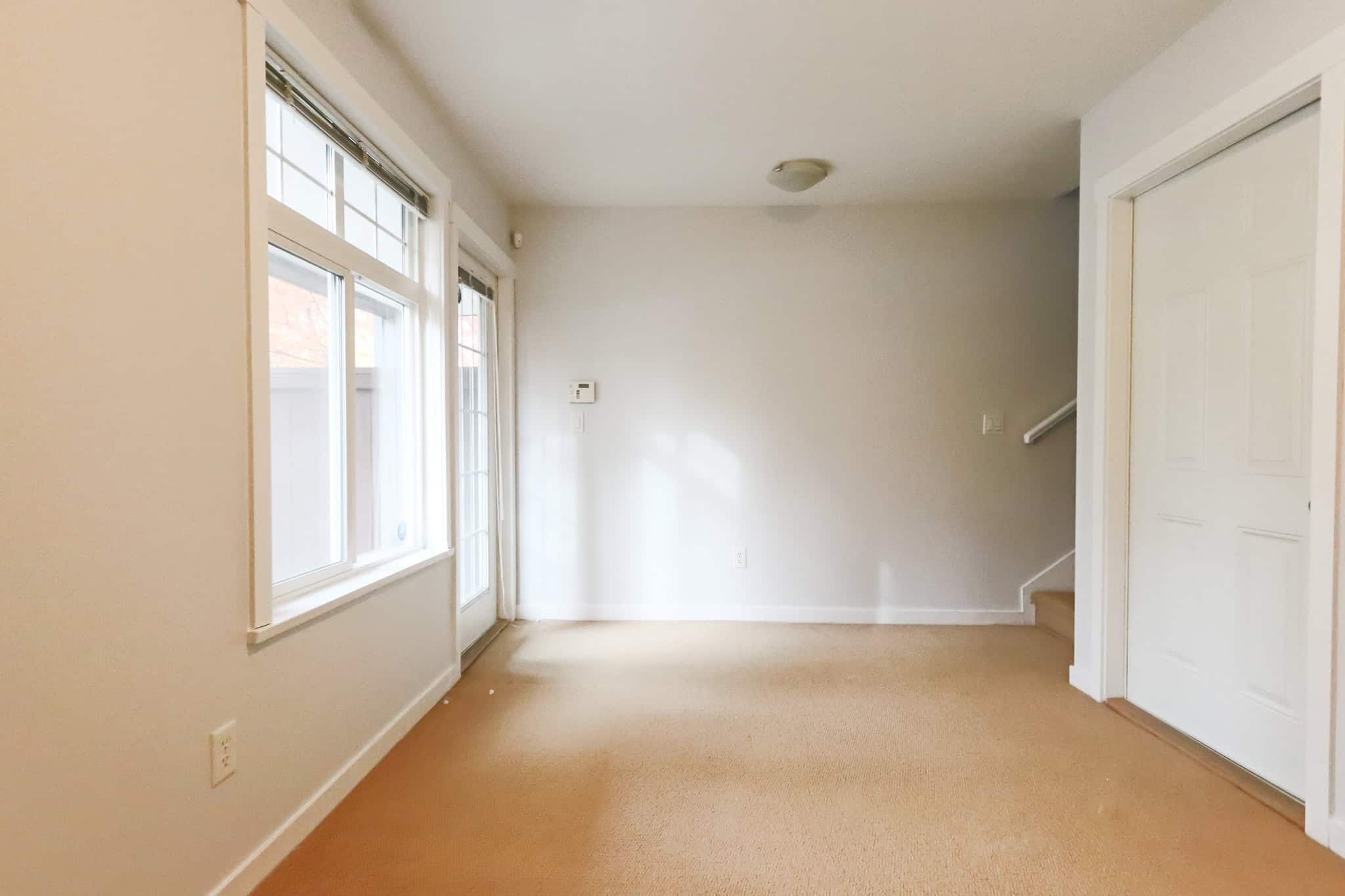 Bright living room with large windows, neutral wall color, beige carpet flooring, and a staircase leading upstairs, ideal for comfortable family living.