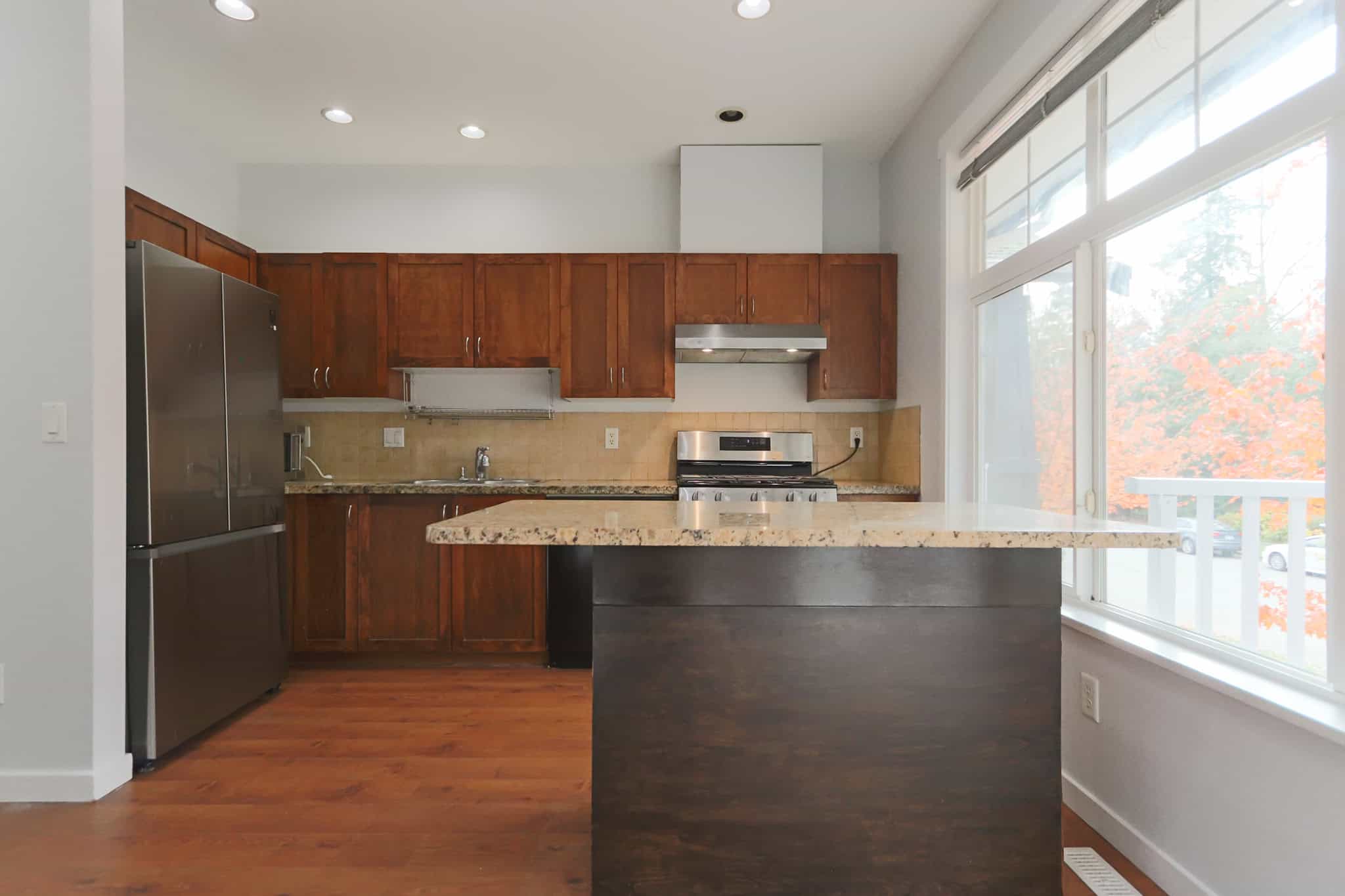 Stainless steel refrigerator and modern kitchen with wood cabinetry, granite countertops, and large windows providing natural light in a cozy home setting.