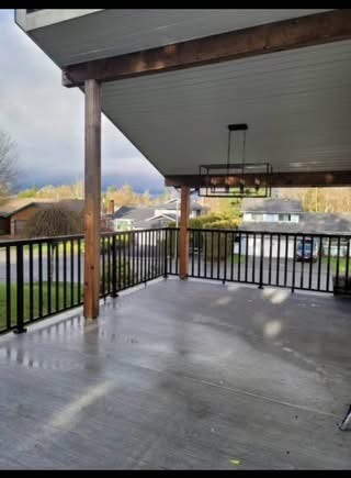 Spacious covered balcony or deck with wooden beams and railing, overlooking a suburban neighbourhood with trees and houses in the distance.