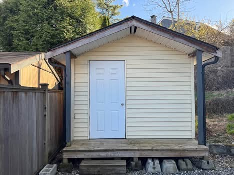 Small backyard shed with white siding and a blue door, elevated on a wooden platform, surrounded by a fence and greenery, ideal for storage or gardening tools.