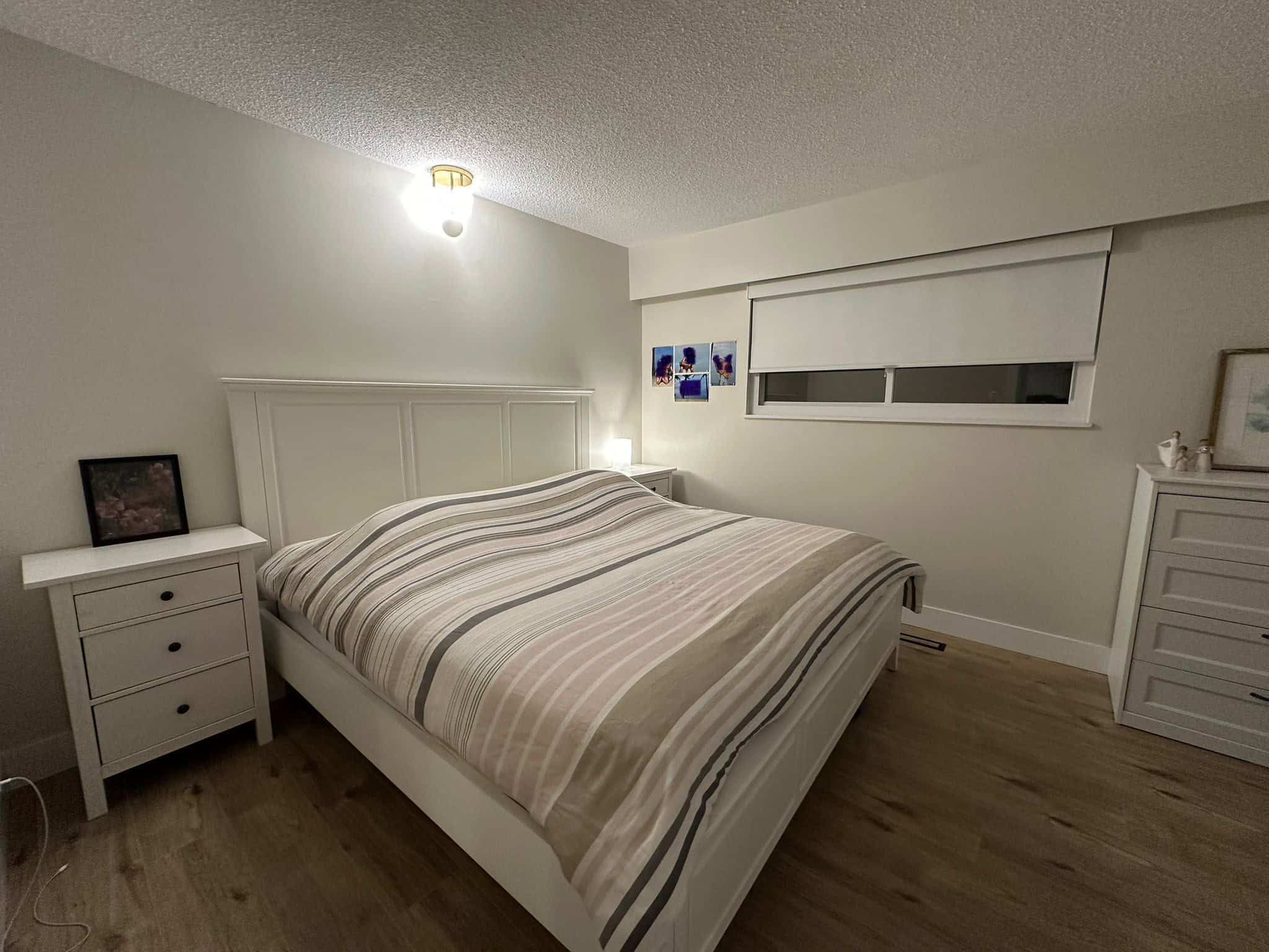 Bright and cozy bedroom featuring a white bed frame, matching nightstands, and a large window with white blinds, perfect for a comfortable home in Vancouver, BC.