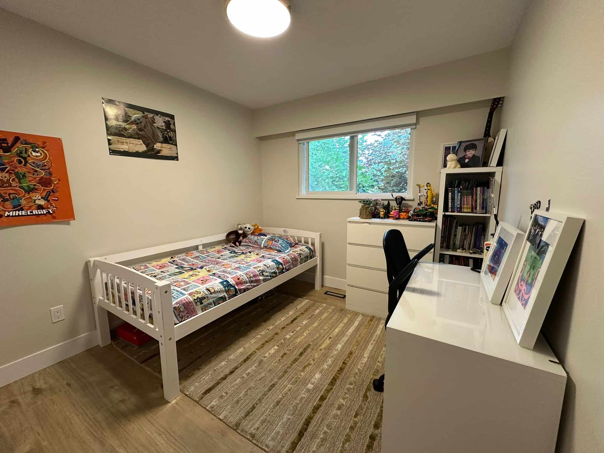 Bright child's bedroom with single white bed, colourful bedding, toys, window view of greenery, and a white desk with framed artwork, perfect for a family home in Vancouver Island.