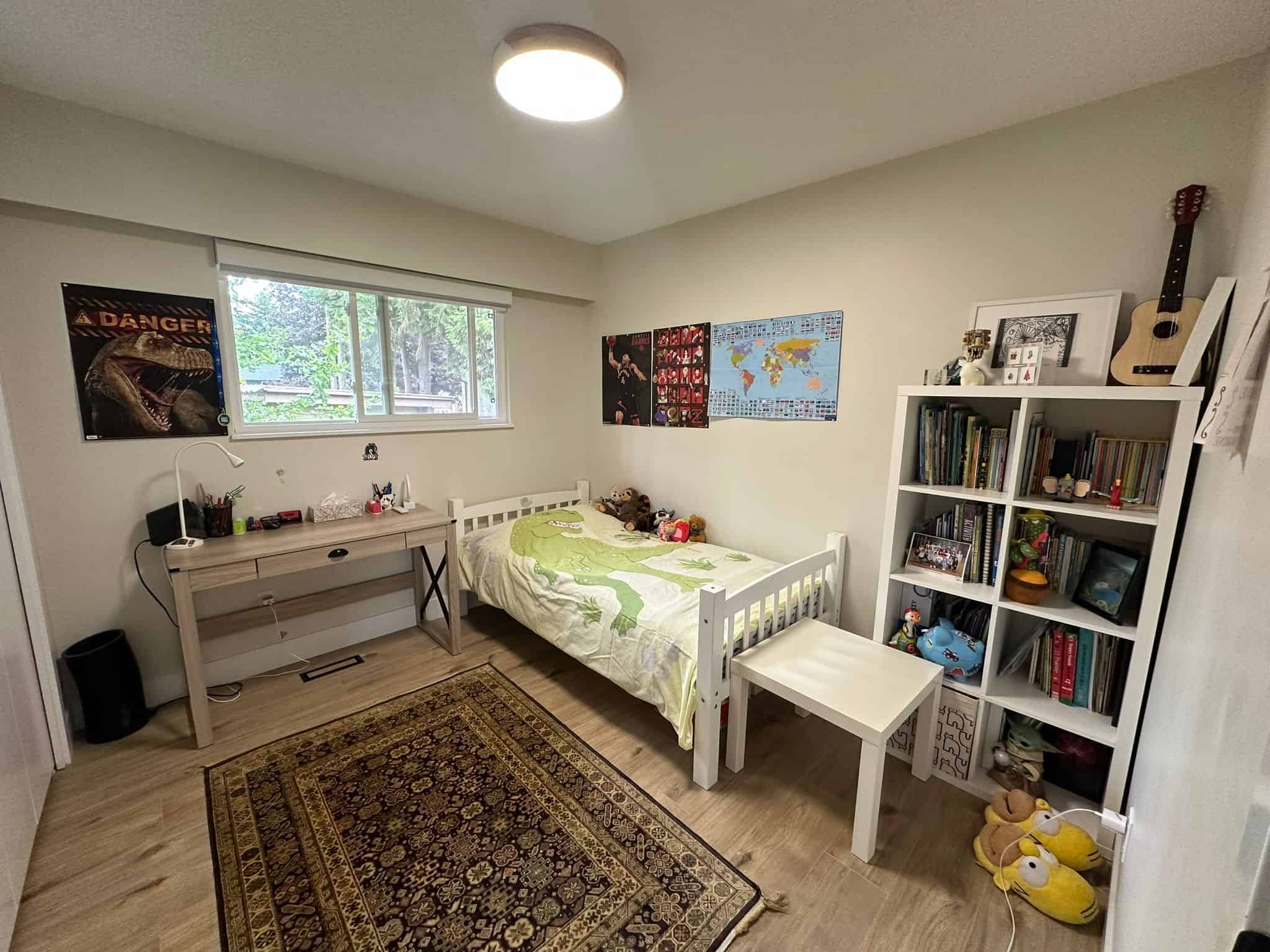 Bright children's bedroom with a single bed, large window, and wooden flooring, featuring a desk, bookshelf filled with books and toys, and colorful decor in a modern home near Orca Realty Inc. in BC.