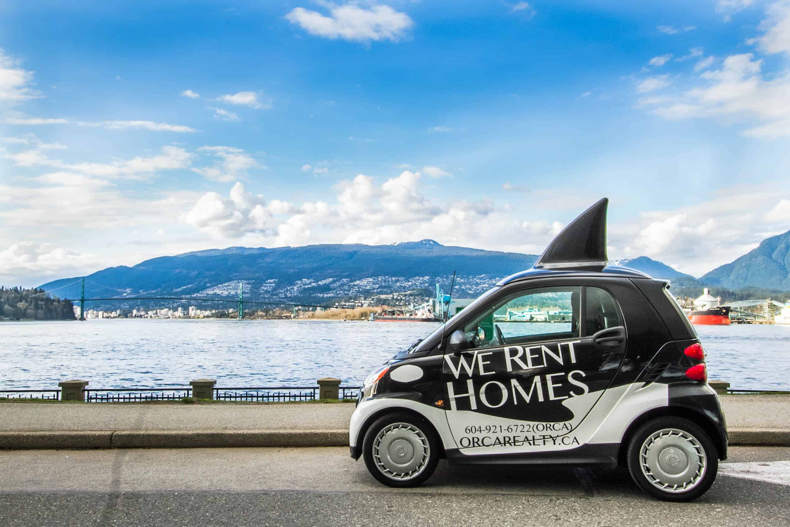 1. Compact electric car with "We Rent Homes" signage parked near waterfront with mountains and cityscape in the background, promoting Orca Realty's home rental services in Vancouver.