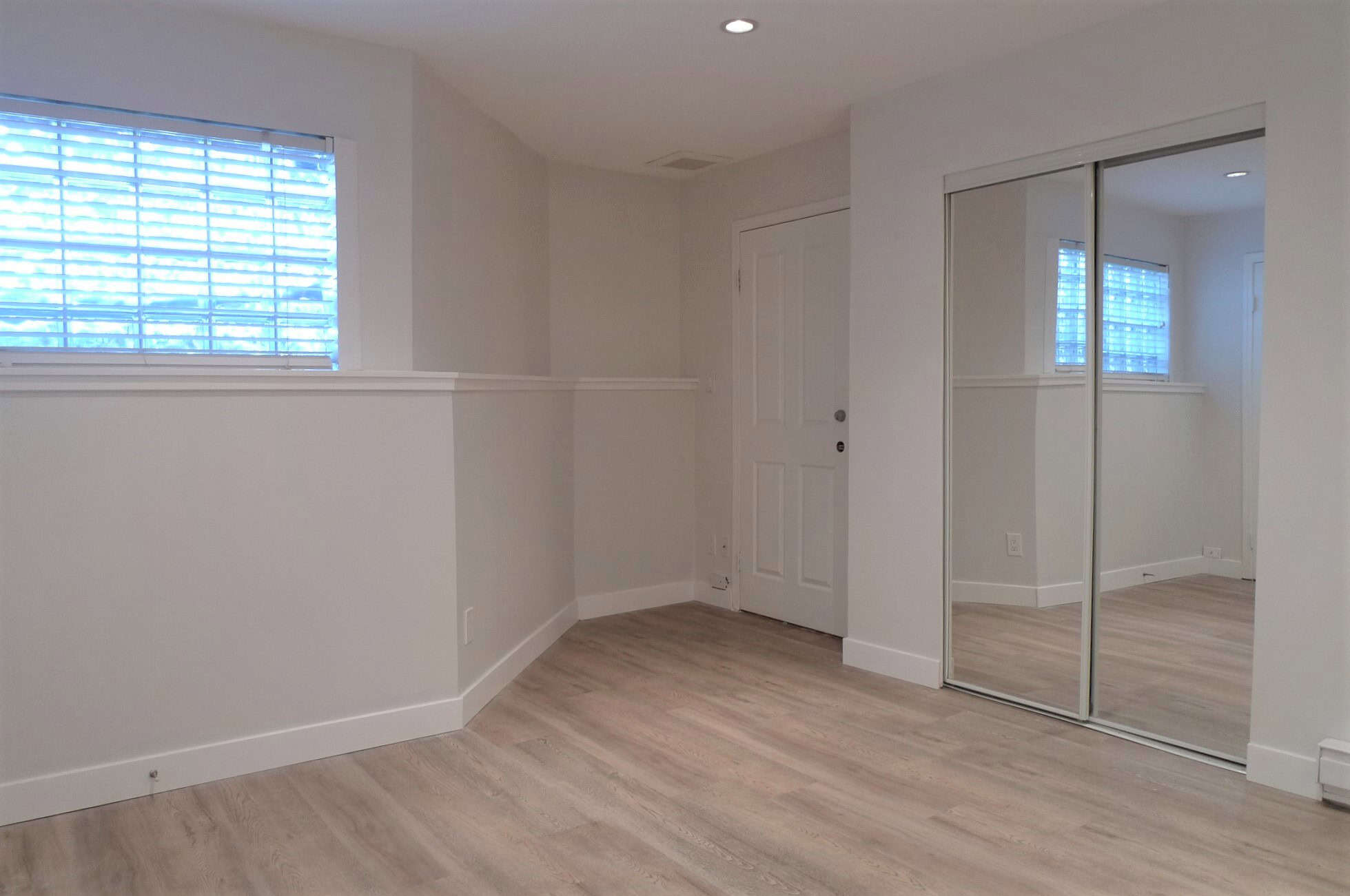 Bright, modern bedroom with white walls, light wood flooring, a window with blinds, and a large mirrored closet, ideal for stylish Vancouver real estate and homebuyers seeking sleek interior design.