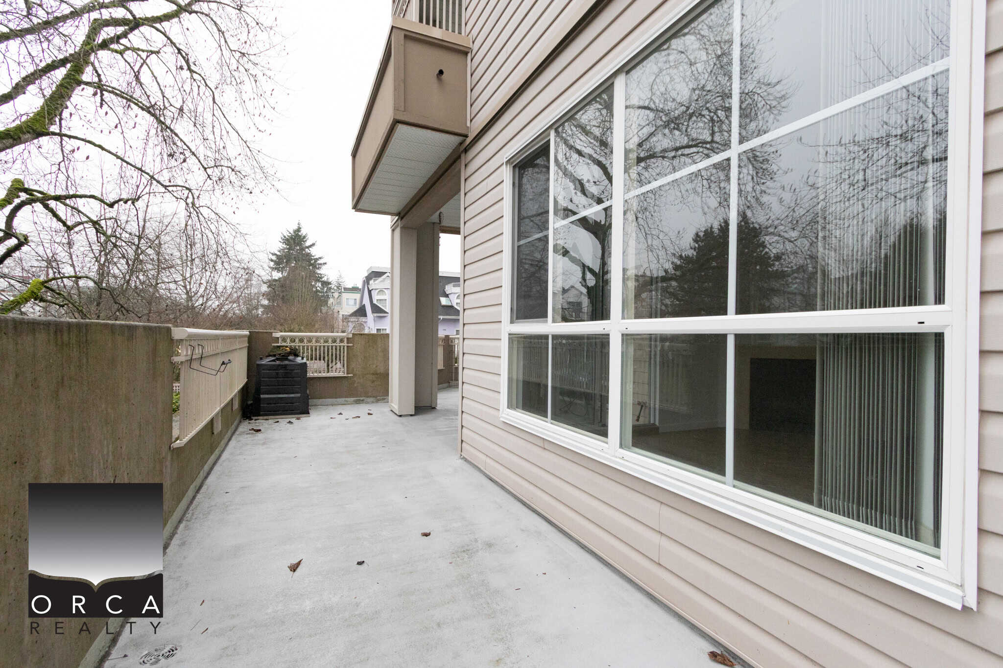 Spacious outdoor balcony with concrete flooring, white railing, and large windows offering natural light, in a modern residential property listed by Orca Realty Inc.