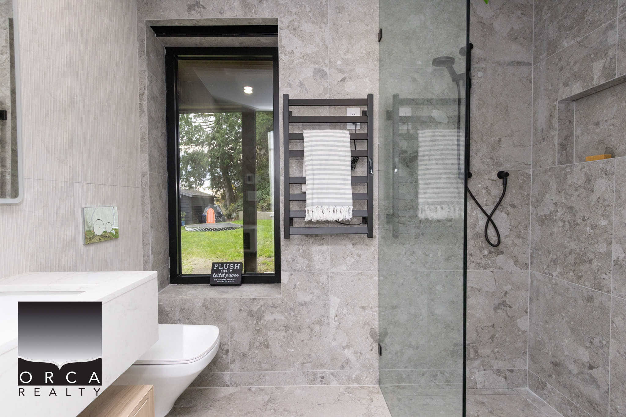 Modern bathroom with walk-in shower featuring grey stone tiles, a sleek black towel rack, and a window overlooking a lush backyard, exemplifying contemporary home design by Orca Realty Inc.