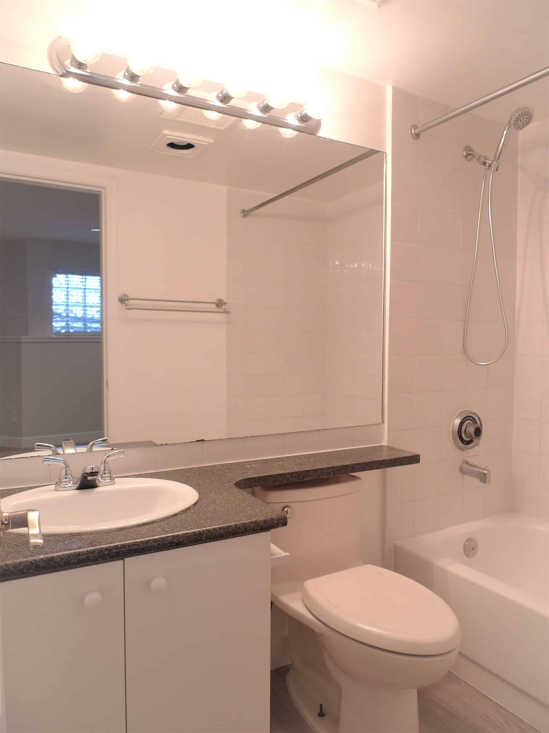 Bright bathroom with white tiles, vanity with dark countertop, large mirror, bathtub with showerhead, toilet, and modern lighting - ideal for Vancouver real estate listings.