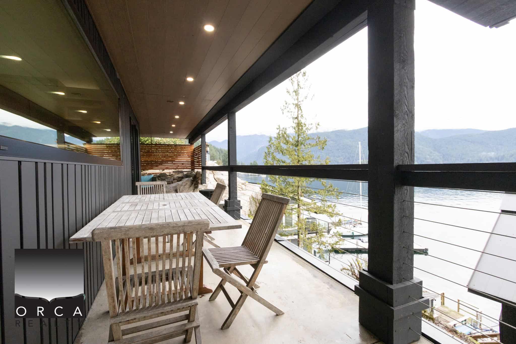 Stunning waterfront balcony with mountain views, outdoor wooden dining table, and modern black railing, perfect for relaxing or entertaining in BC's scenic coastal location.