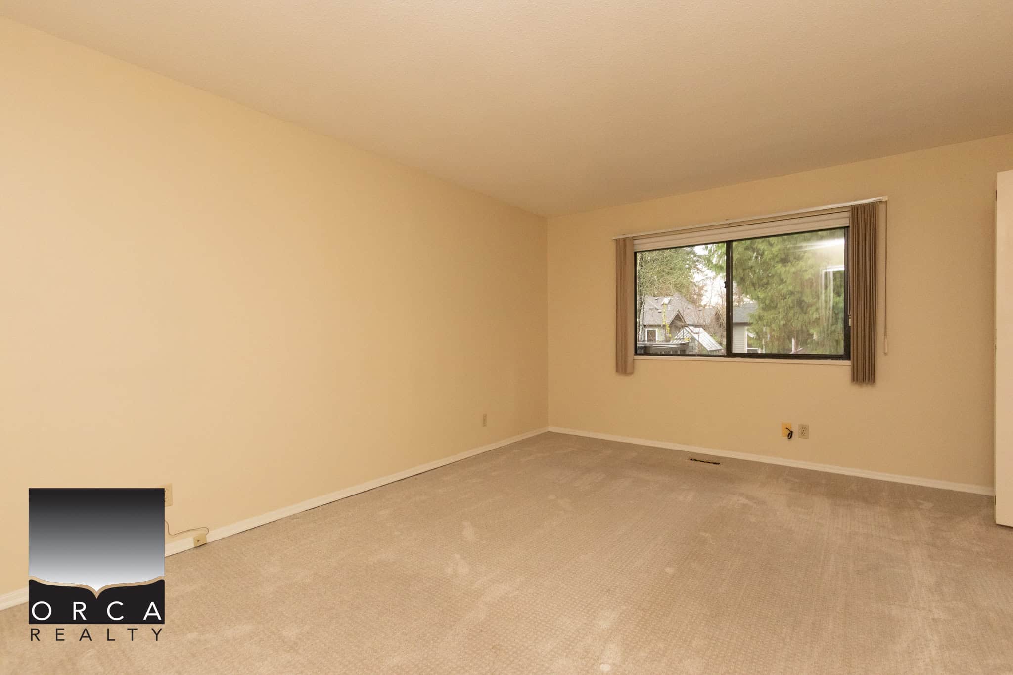 Bright empty living room with large window, beige walls, and carpeted flooring, ideal for homebuyers seeking a spacious and well-lit Vancouver Island property from Orca Realty Inc.