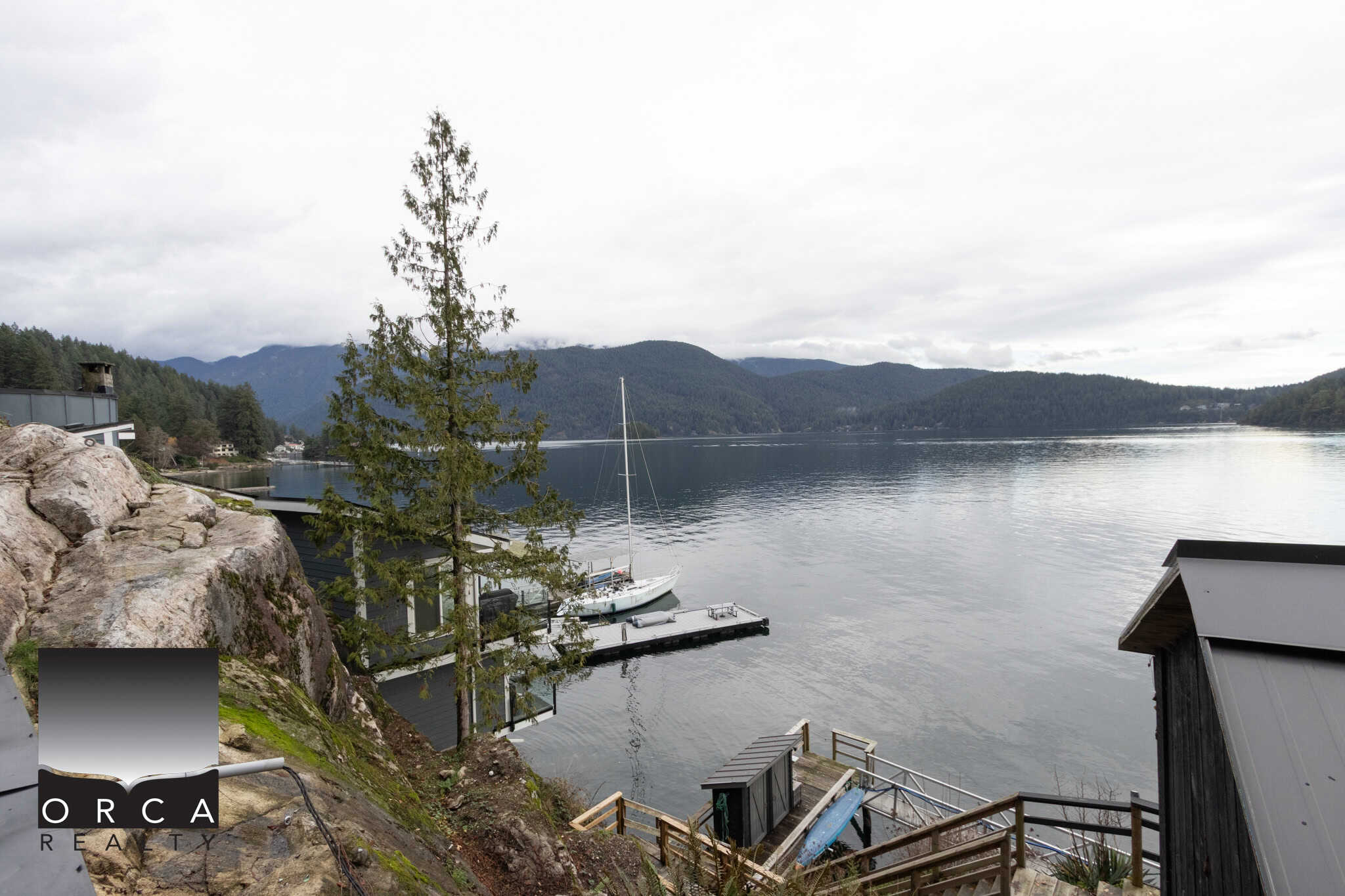 Serene waterfront view of a lake with a sailboat docked at a modern boathouse, surrounded by lush mountains and forest, showcasing exclusive real estate opportunities on Orca Island.
